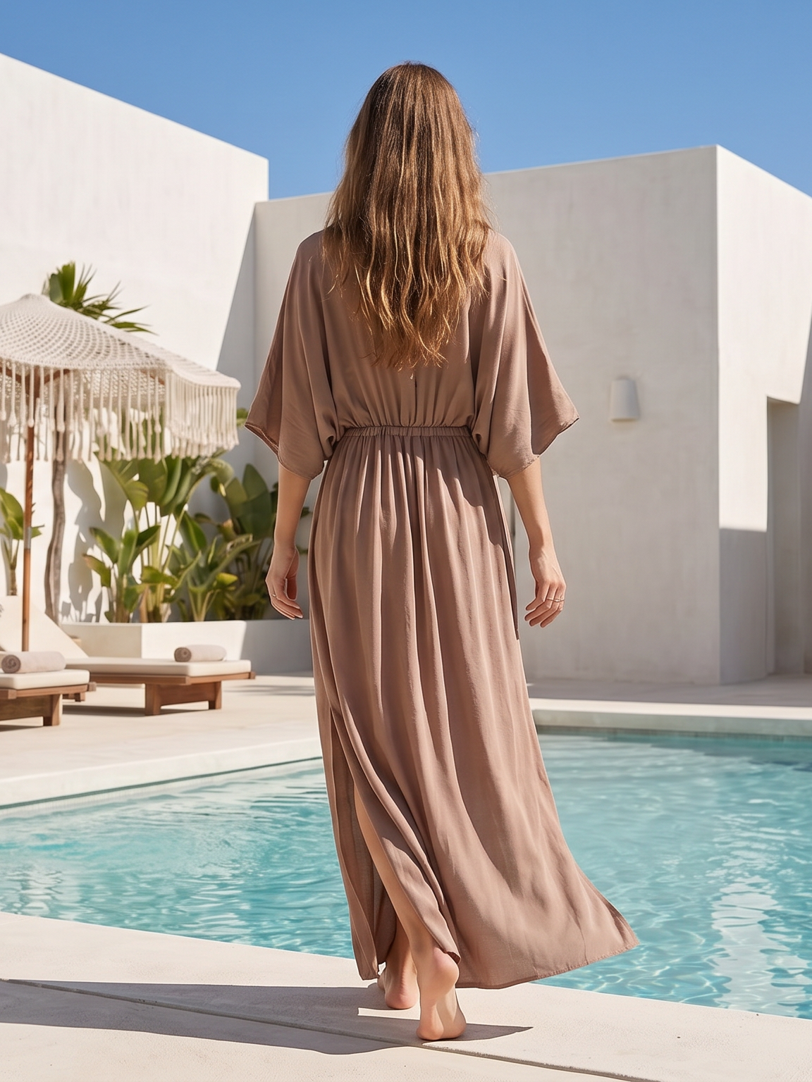 Woman in a long taupe caftan dress walking by a pool with a white building and greenery in the background.