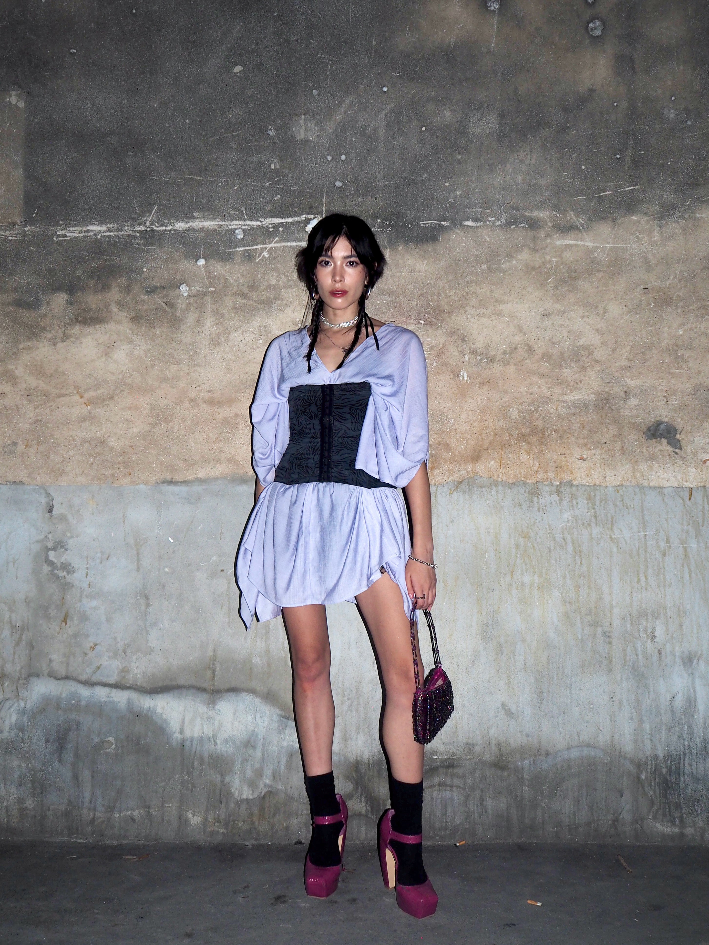 A woman stands on wearing a flowing lavender beach cover-up mini kaftan dress, with ocean waves and old wall in the background.