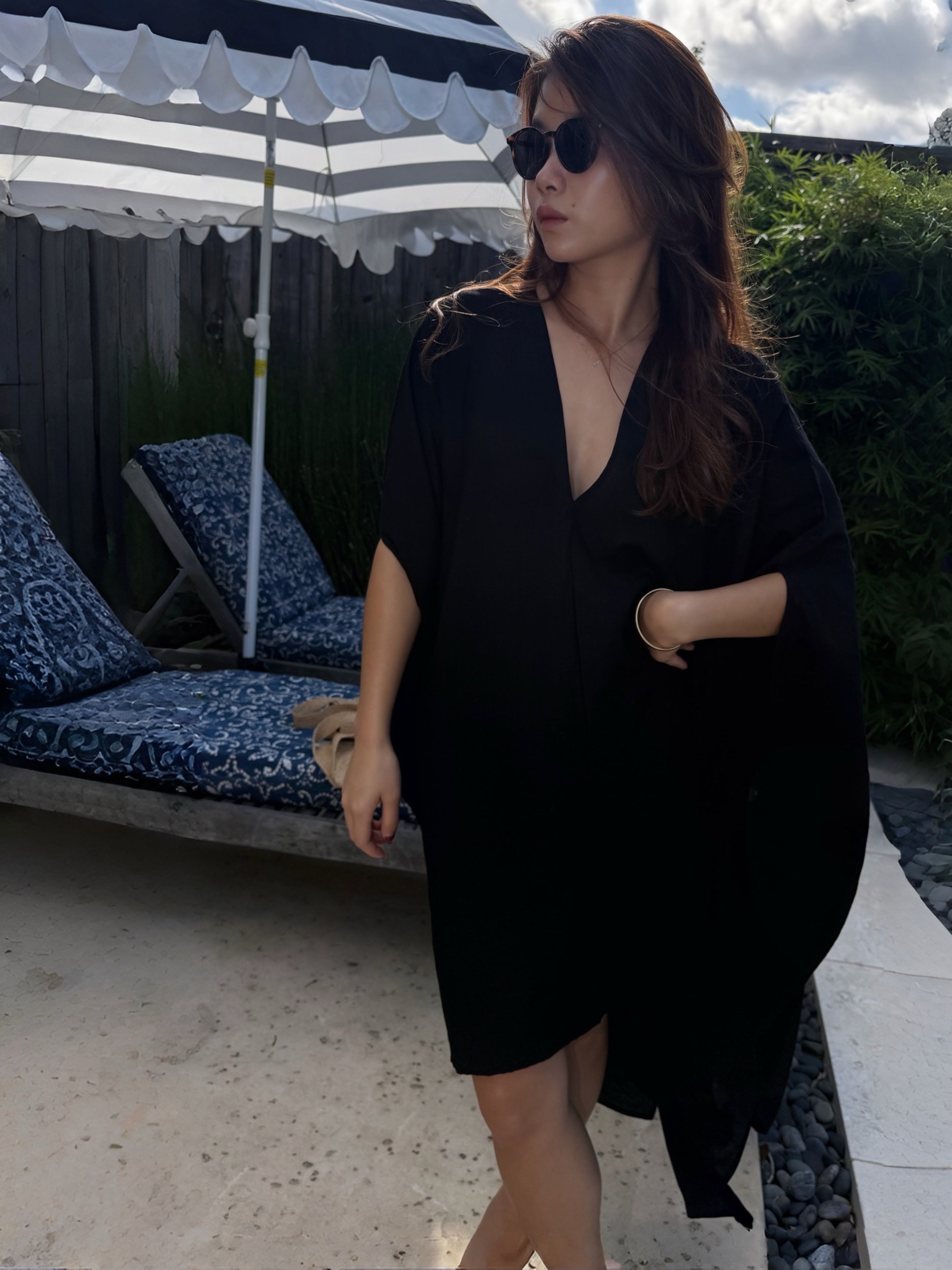 A woman wearing a Black Mini Kaftan Cover Up, oversized beach cover up and petite mini dress with deep V neckline for resort wear and poolside vacation outfits stands by a swimming pool under a white patio umbrella, with green foliage in the background.