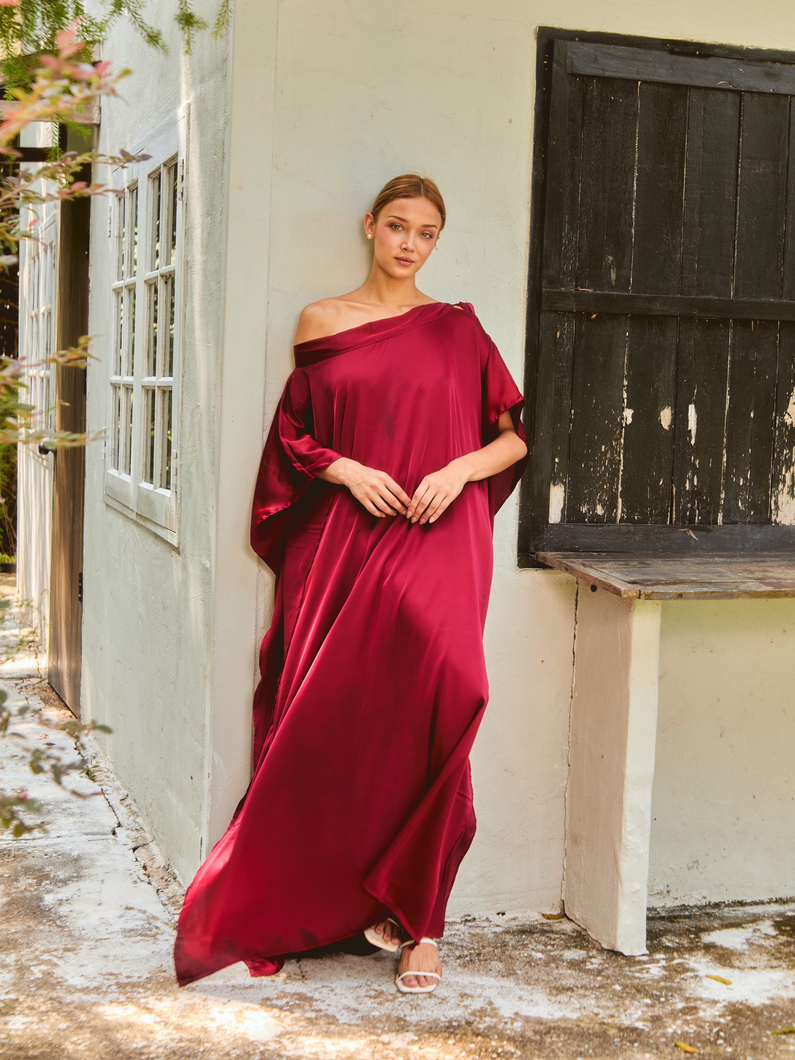 Woman in a burgundy off-shoulder dress standing outdoors near a white wall. The Helena Red Burgundy Kaftan by Coco De Chom features an elegant, free-flowing silhouette with tie details, made from silky eco-friendly satin fabric. Perfect for resort wear, beach vacations, and effortless boho luxury.