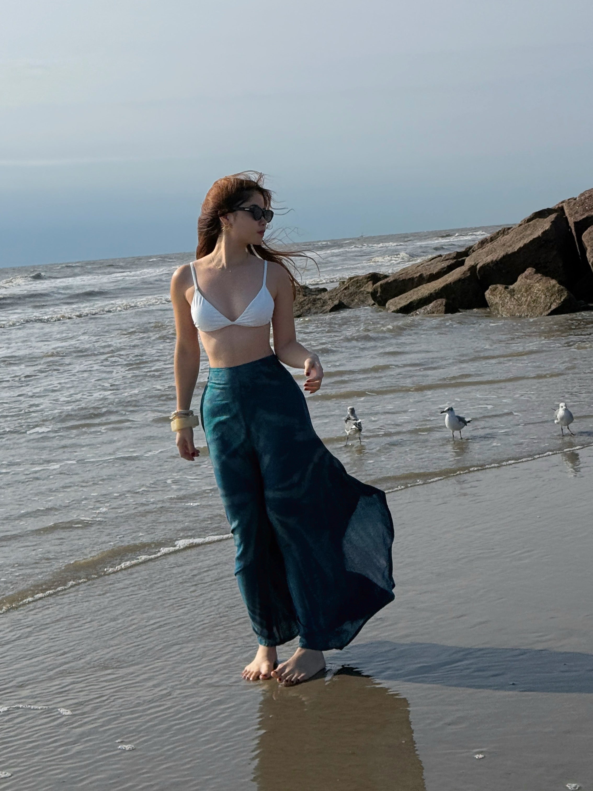 Woman wearing a white top and teal tie dye boho pants standing against a white wall with sand at the bottom, perfect beach outfit.