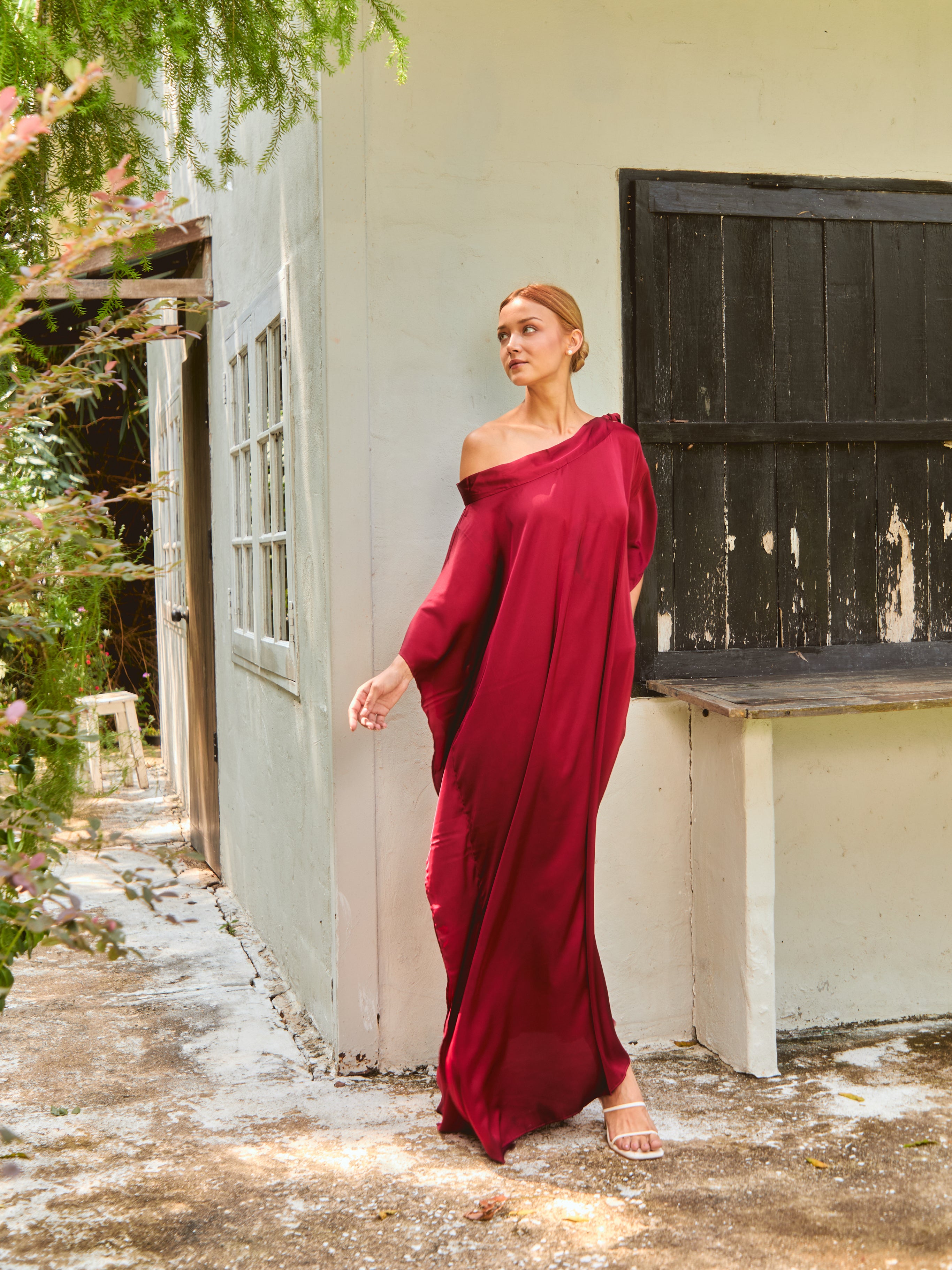 Woman in a burgundy off-shoulder dress standing outdoors near a white wall. The Helena Red Burgundy Kaftan by Coco De Chom features an elegant, free-flowing silhouette with tie details, made from silky eco-friendly satin fabric. Perfect for resort wear, beach vacations, and effortless boho luxury.