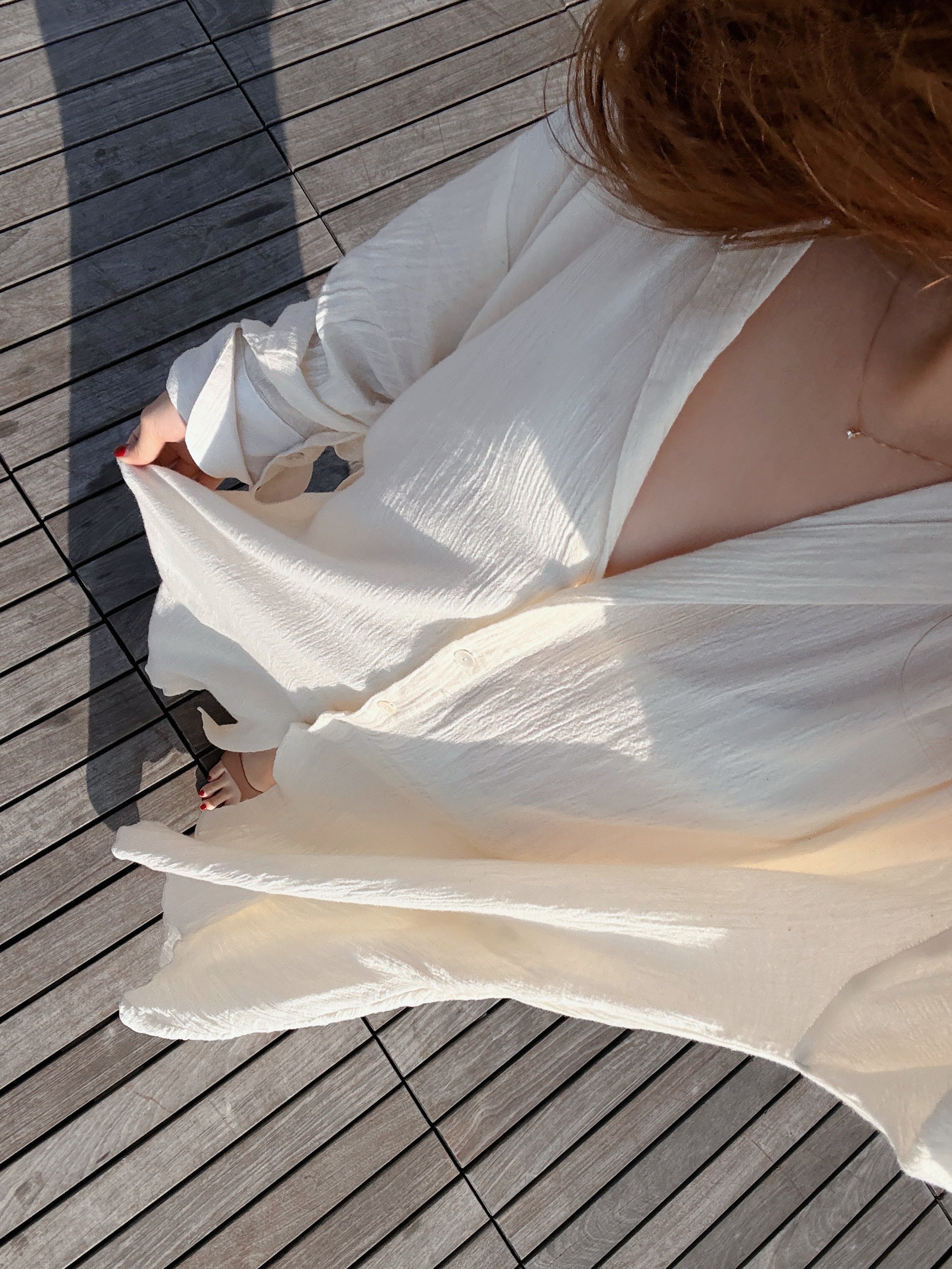 Person wearing a white natural organic cotton button down shirt dress, relaxed boho maxi beach cover up for resort wear on a wooden deck