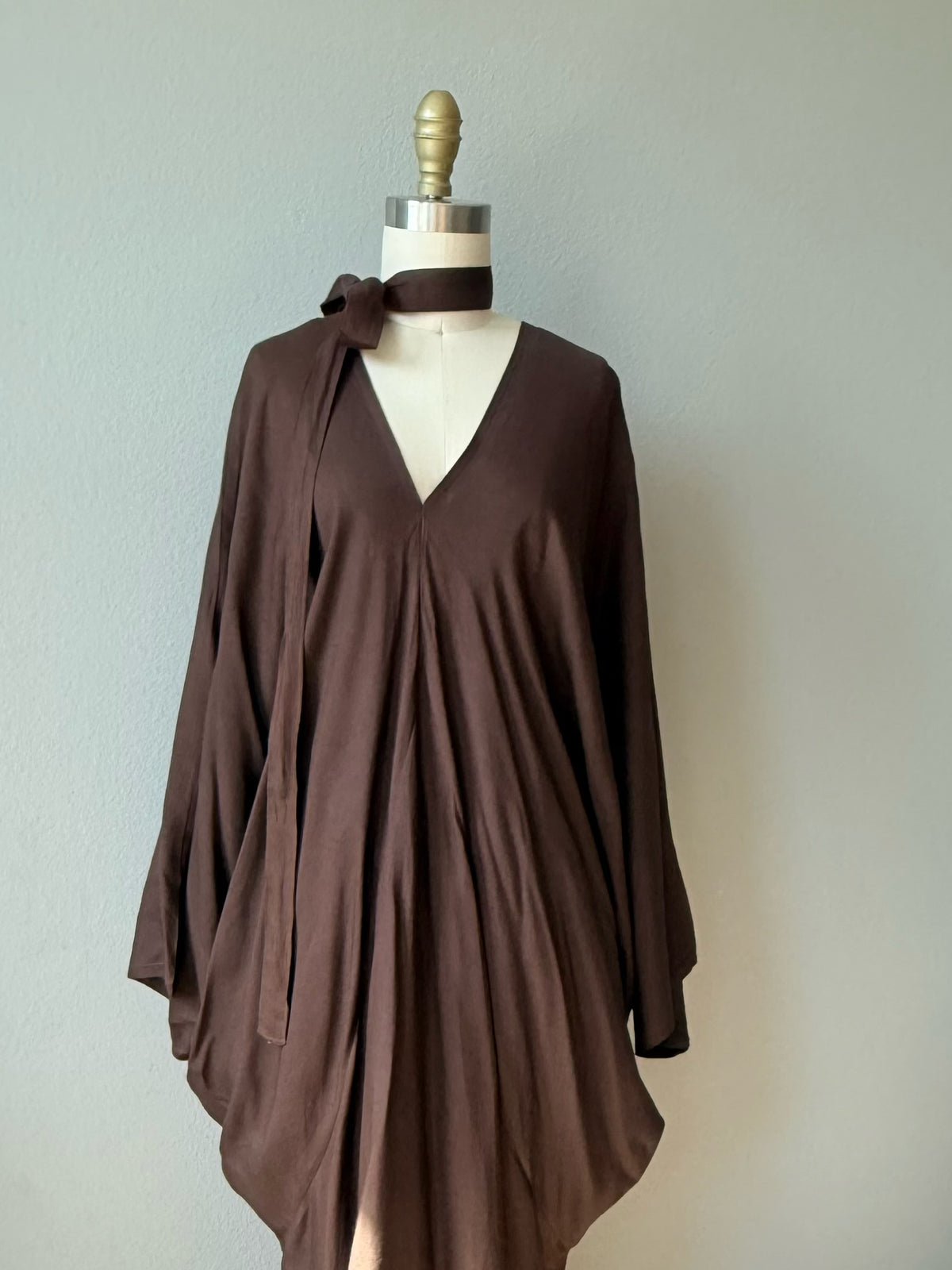 A dark brown kaftan dress oversized style with a V-neckline and tie detail at the collar, displayed on a mannequin against a plain light gray wall.