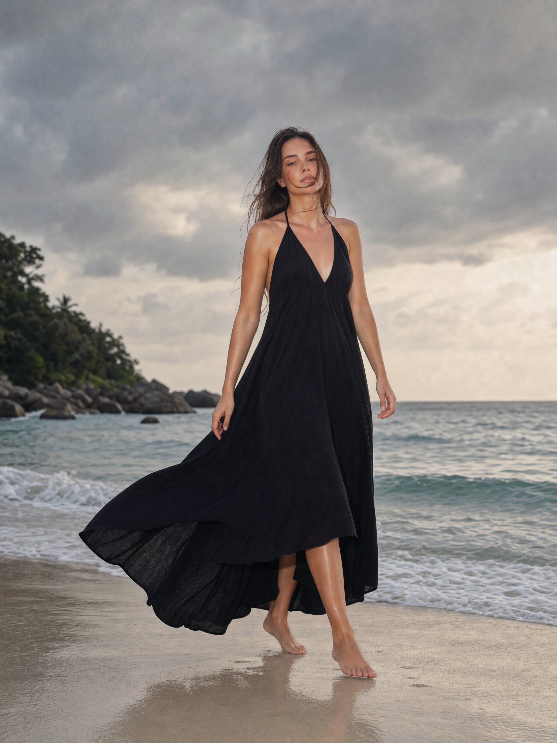 A woman wearing a flowing black halter maxi dress walks barefoot on a sandy beach with ocean waves and a cloudy sky in the background.