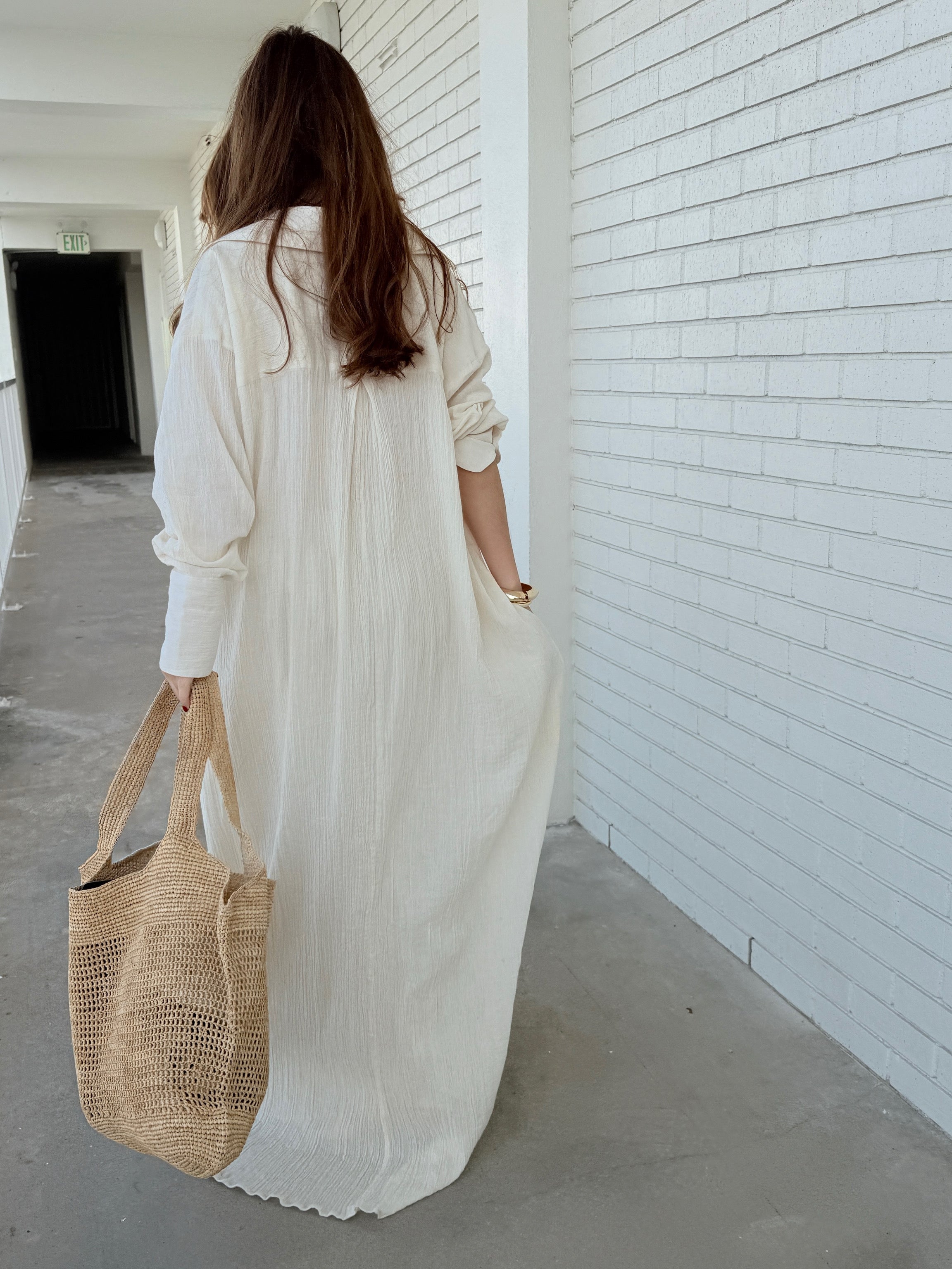 Woman in a long white cotton button down shirt dress, relaxed boho maxi beach cover up for resort wear holding a woven bag against a white wall.