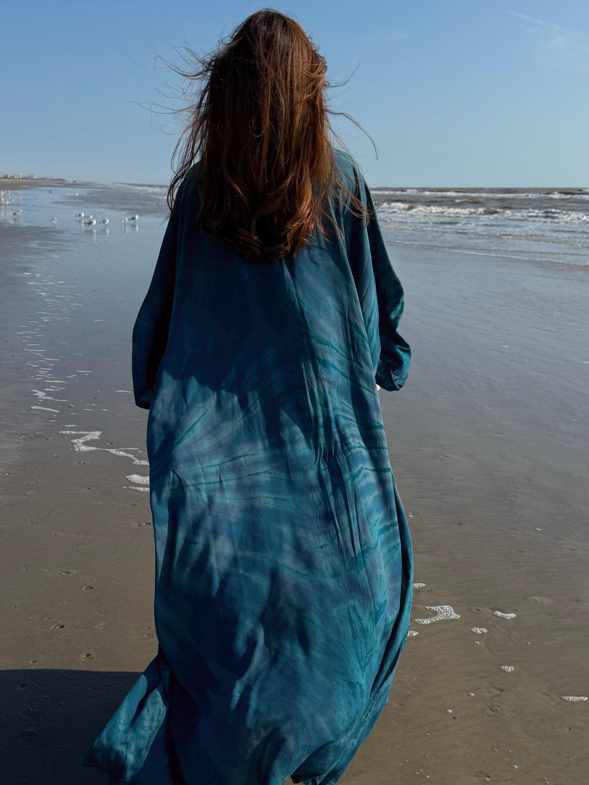 Woman wearing a long teal seashell tie dye kimono robe over a white bikini on the beach – boho beach cover up and vacation resort wear by Coco De Chom.