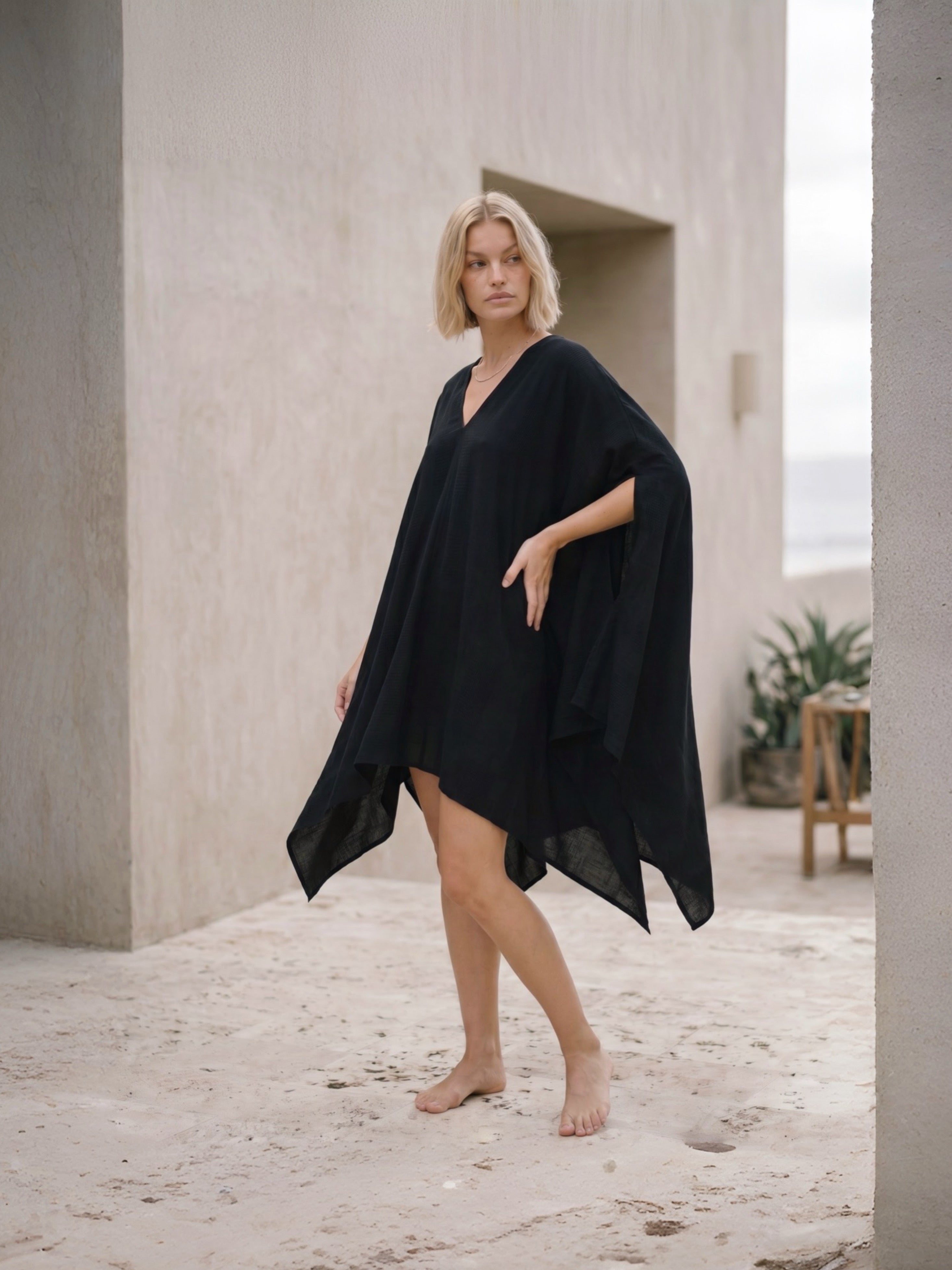 A woman modeling a black, lightweight, flowy kaftan style cover-up dress in an outdoor architectural setting with textured beige walls and a sandy floor. Blac Mini Kaftan Cover Up, oversized beach cover up and petite mini dress with deep V neckline for resort wear and poolside vacation outfits