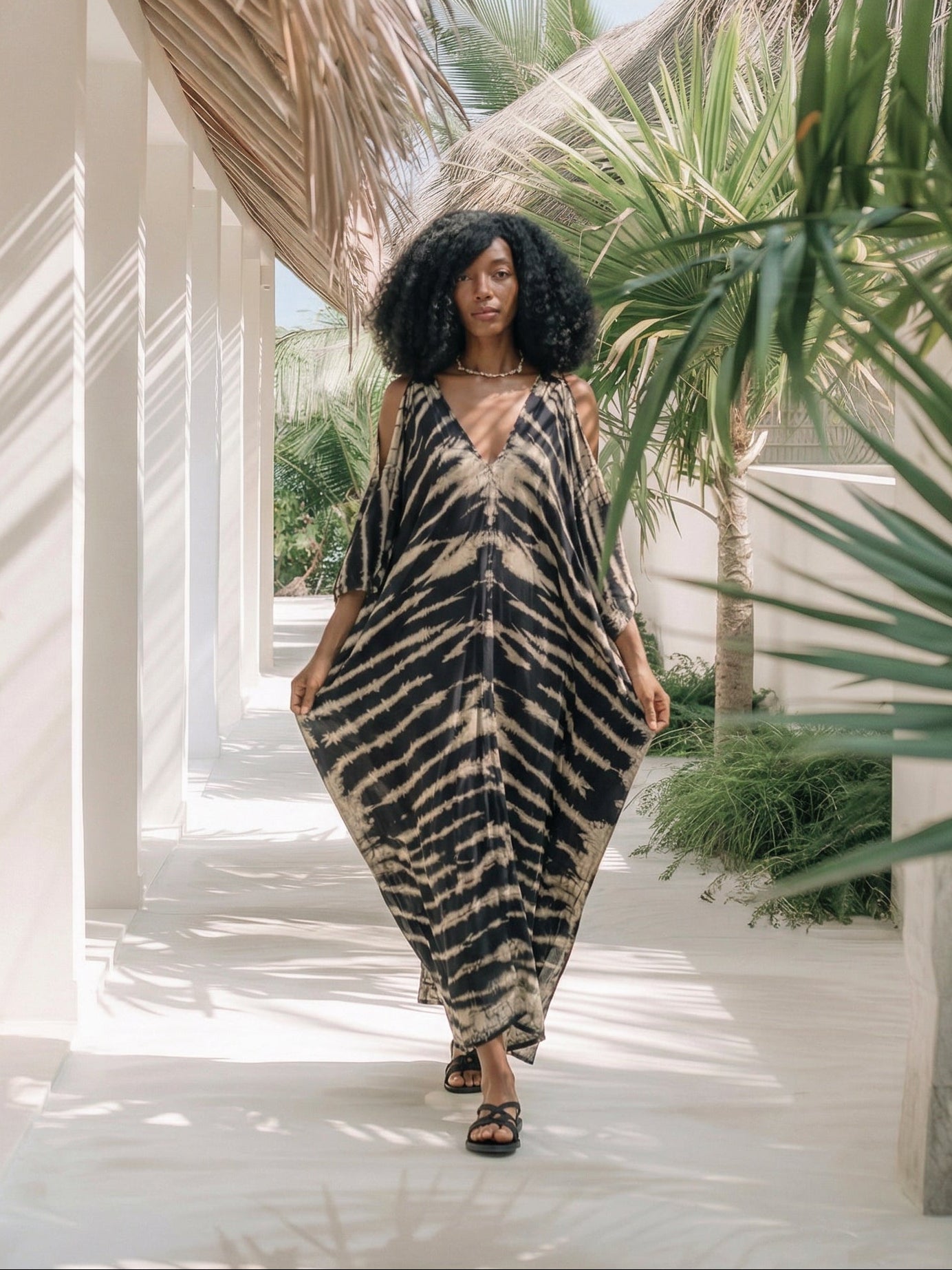 Woman in a tie dye kaftan zebra print in black standing in a sunlit hallway with plants