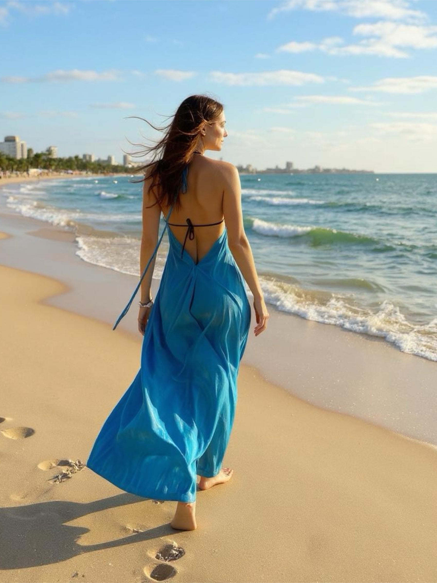 Woman in a blue tie dye dress walking on a beach with a city skyline in the background, perfect for beach dress, resort wear