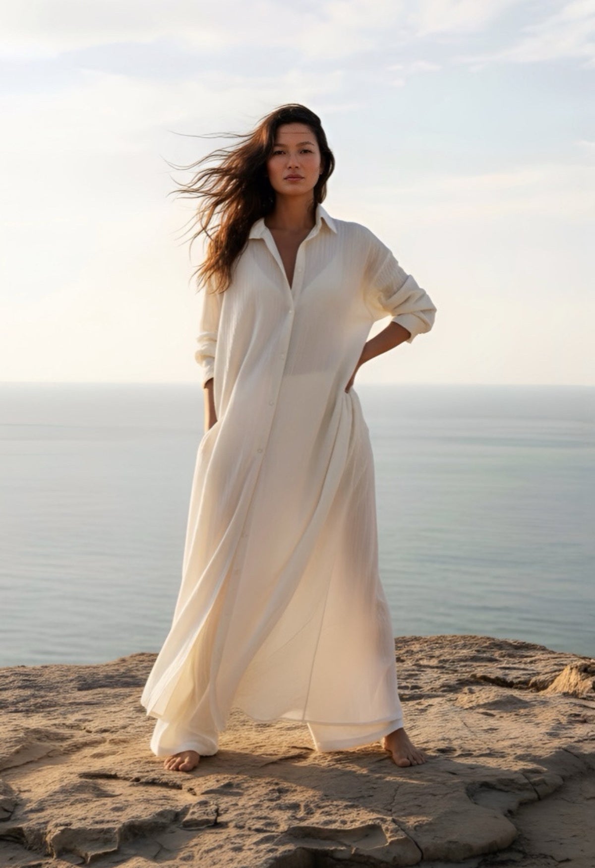 Woman in a white cotton shirt dress standing on a rocky beach with ocean and sky in the background