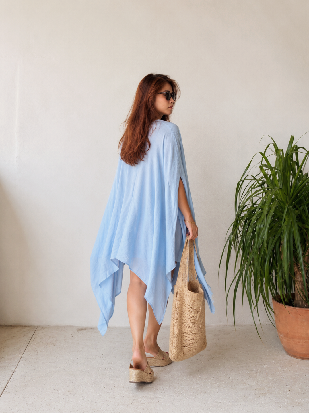 A woman modeling a light blue kaftan dress with a V-neckline and wide draped sleeves, standing against a neutral light gray background. Sky blue Soleil Mini Kaftan Cover Up, lightweight mini kaftan dress and beach cover up styled as a resort top for summer vacation