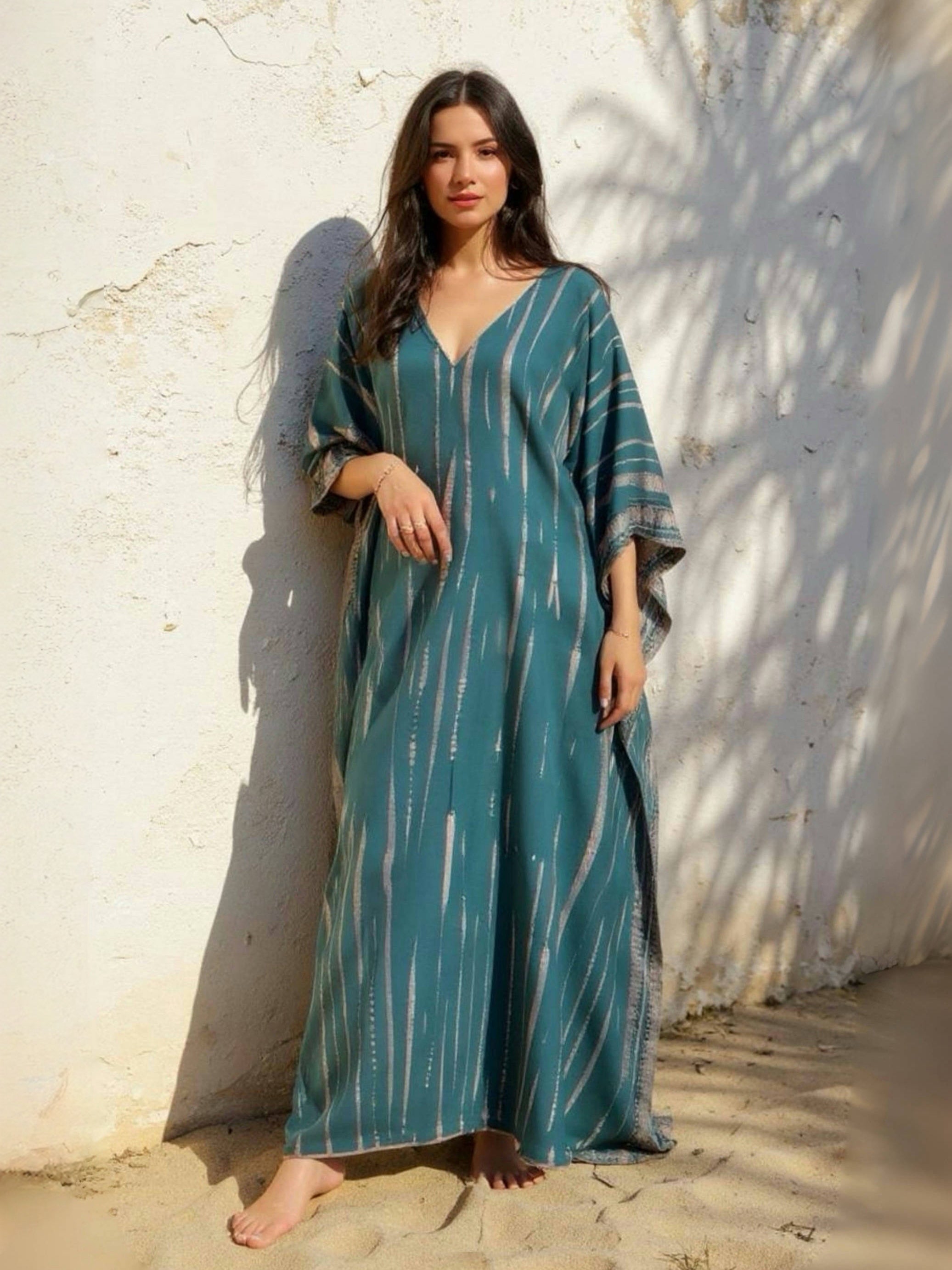 Woman wearing a teal Maxi Kaftan in tie-dye pattern with V-neck and side slits against a textured wall.