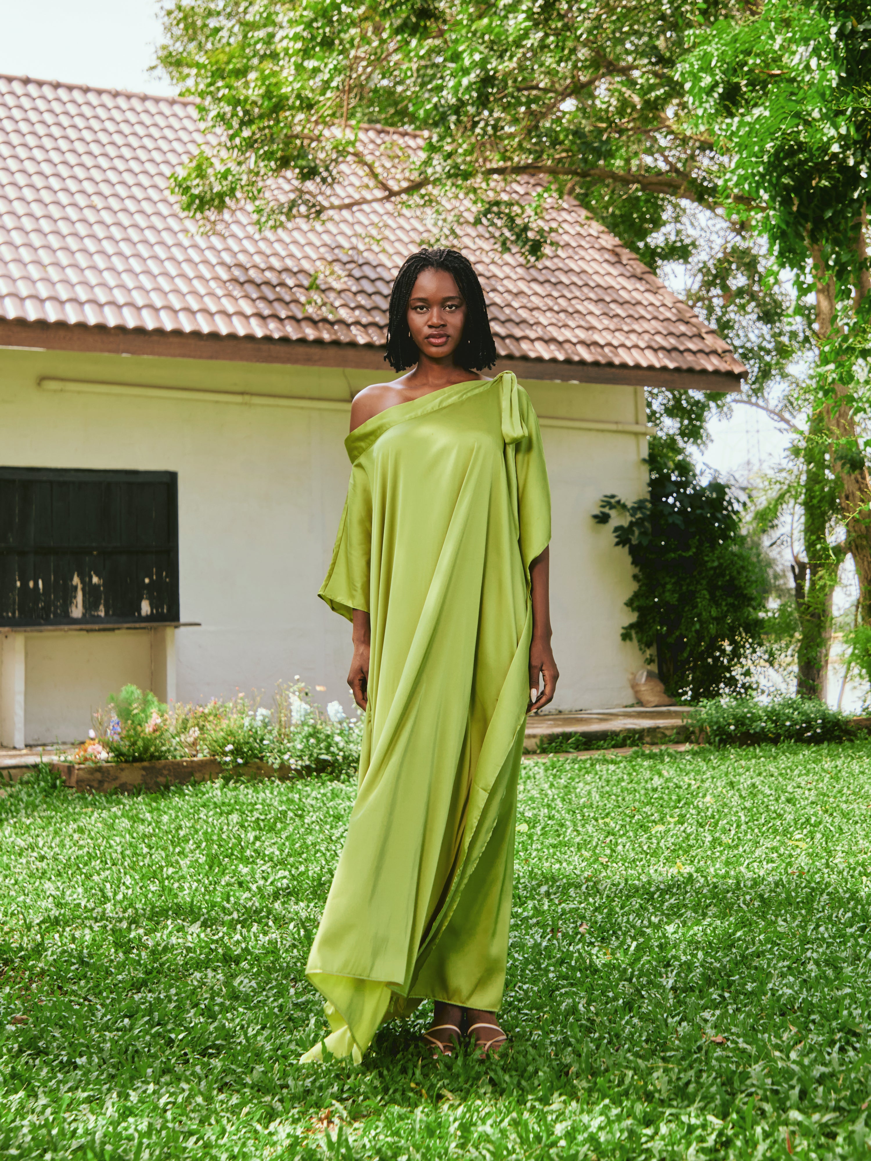 Woman in a lemon green kaftan dress standing on grass with a house and tree in the background. The Helena Lemon Green Kaftan by Coco De Chom is an off-shoulder satin resort dress with a free-flowing silhouette, tie details, and silky eco-friendly fabric — perfect for beach vacations, resort wear, and effortless boho luxury.