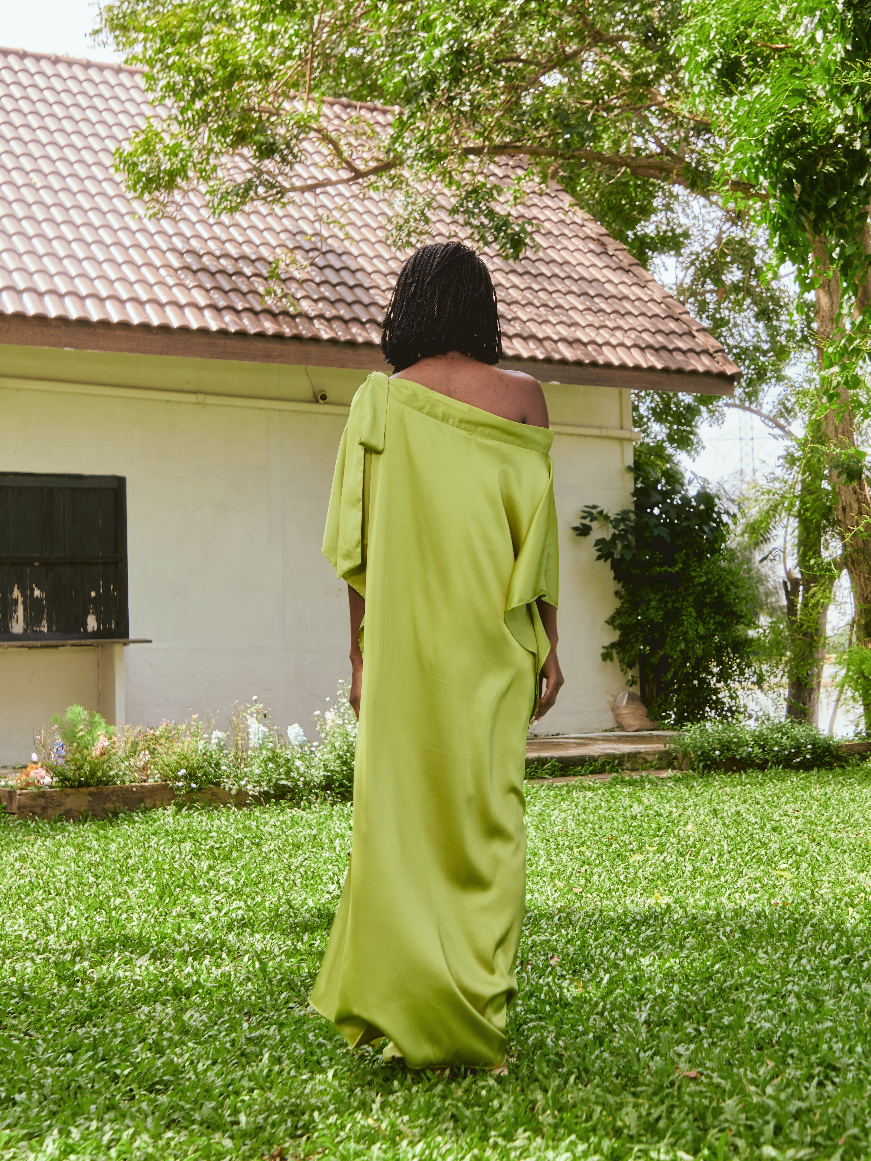 Woman in a lemon green kaftan dress standing on grass with a house and tree in the background. The Helena Lemon Green Kaftan by Coco De Chom is an off-shoulder satin resort dress with a free-flowing silhouette, tie details, and silky eco-friendly fabric — perfect for beach vacations, resort wear, and effortless boho luxury.