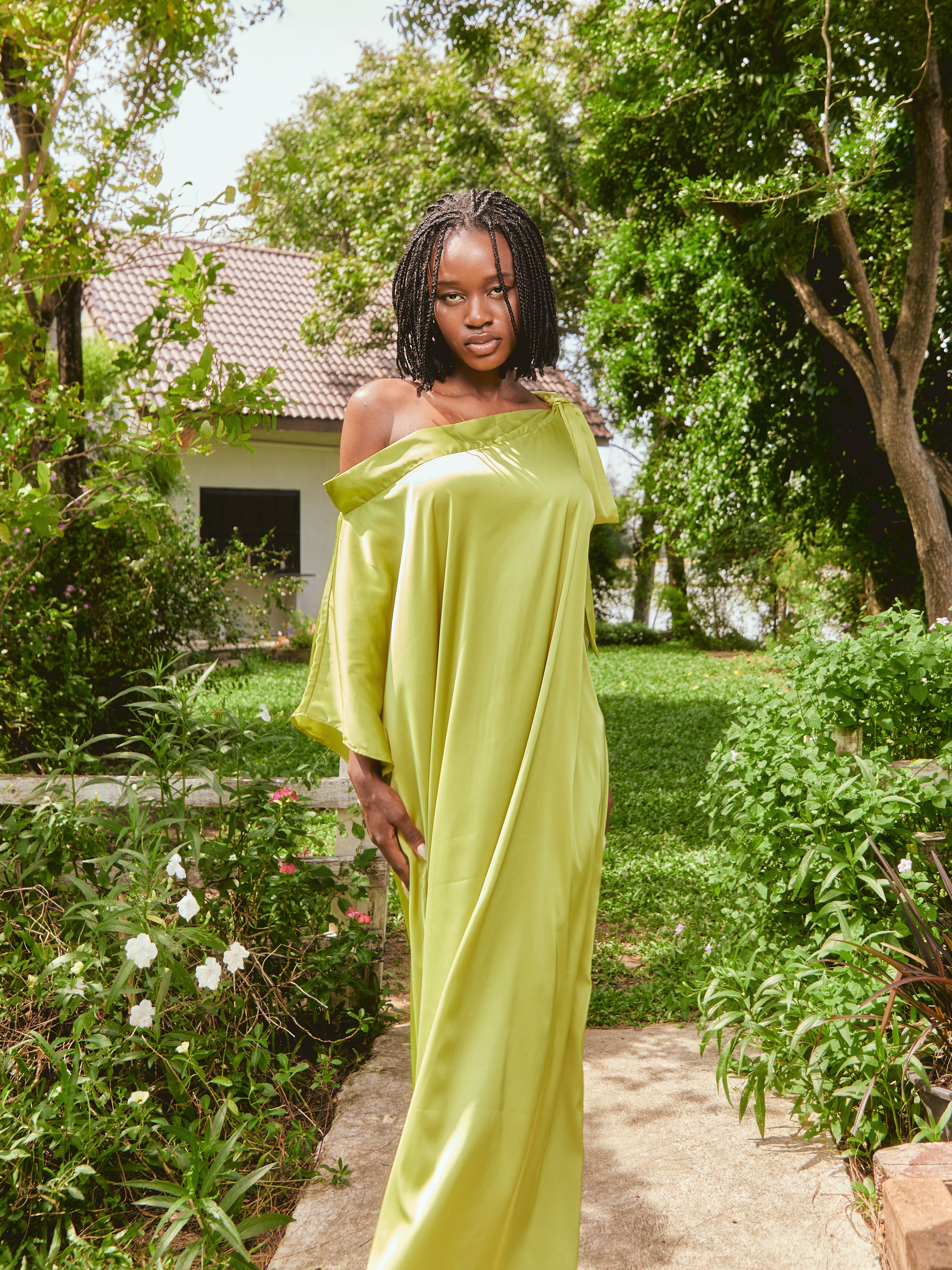 Woman in a lemon green kaftan dress standing on grass with a house and tree in the background. The Helena Lemon Green Kaftan by Coco De Chom is an off-shoulder satin resort dress with a free-flowing silhouette, tie details, and silky eco-friendly fabric — perfect for beach vacations, resort wear, and effortless boho luxury.