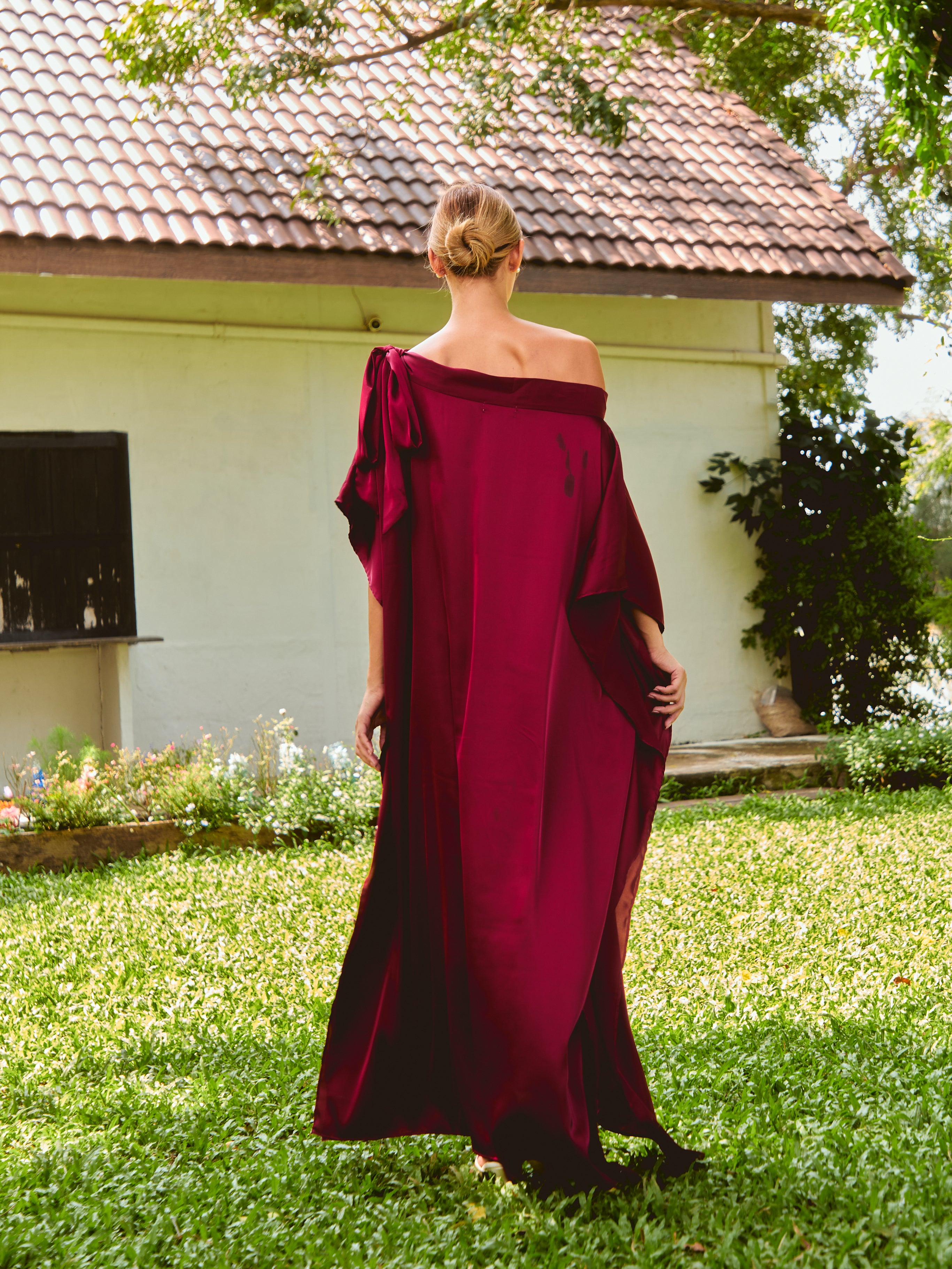 Woman wearing a burgundy off-shoulder dress standing outdoors. The Helena Red Burgundy Kaftan by Coco De Chom features an elegant, free-flowing silhouette with tie details, made from silky eco-friendly satin fabric. Perfect for resort wear, beach vacations, and effortless boho luxury.