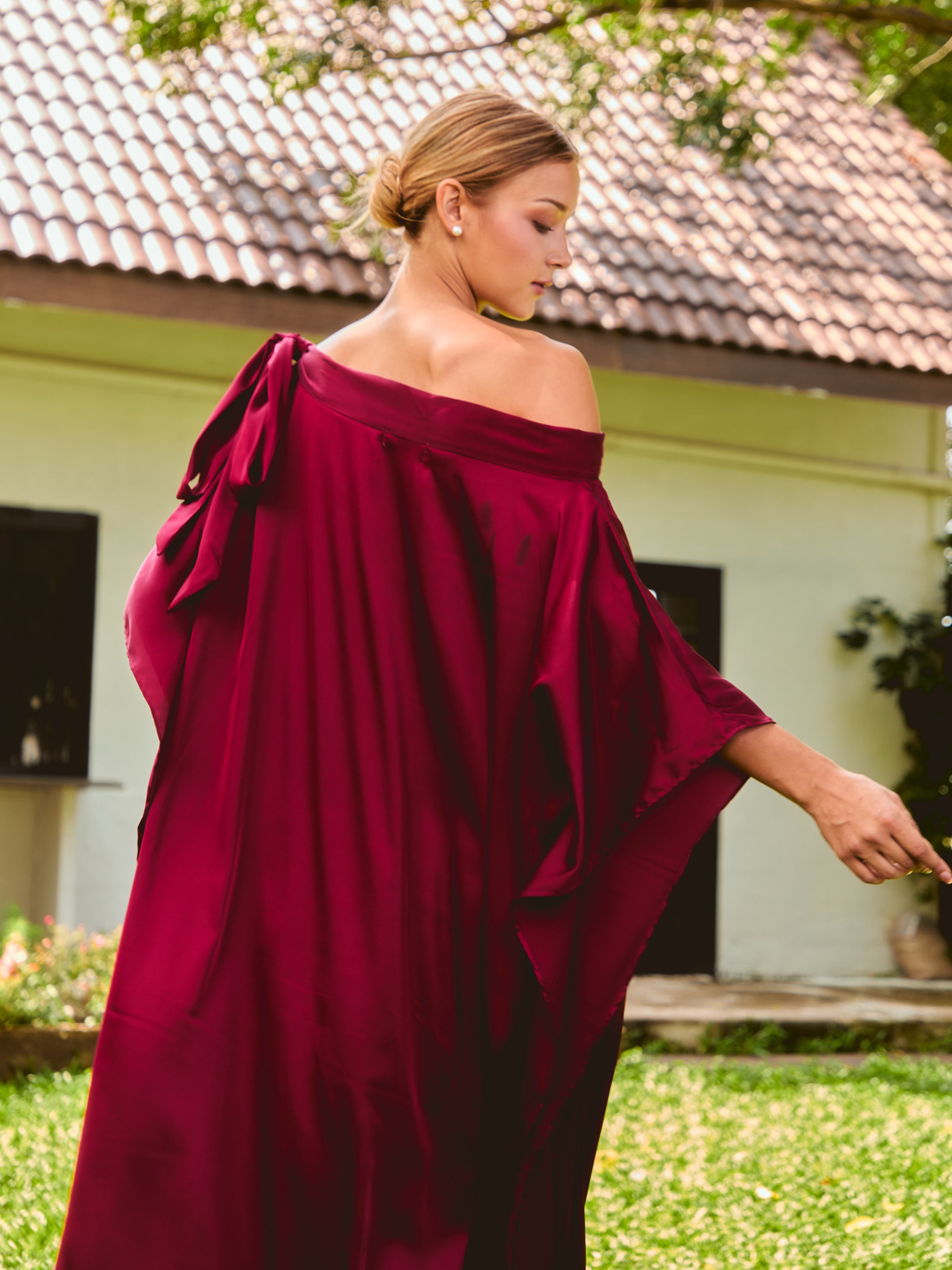 Woman wearing a burgundy off-shoulder dress standing outdoors. The Helena Red Burgundy Kaftan by Coco De Chom features an elegant, free-flowing silhouette with tie details, made from silky eco-friendly satin fabric. Perfect for resort wear, beach vacations, and effortless boho luxury.