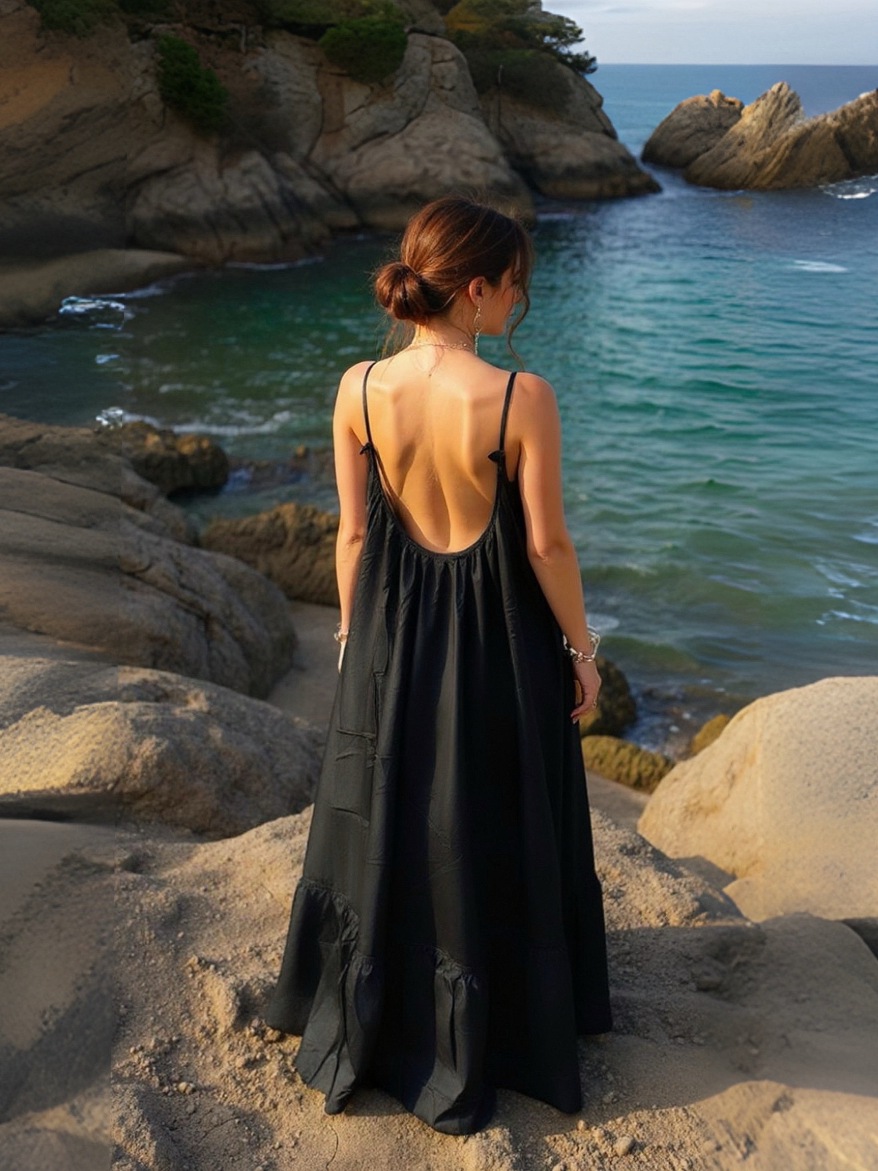 Woman in a hand-dyed black backless maxi dress standing by the sea, holding a glass of wine, wearing boho resortwear by Coco de Chom