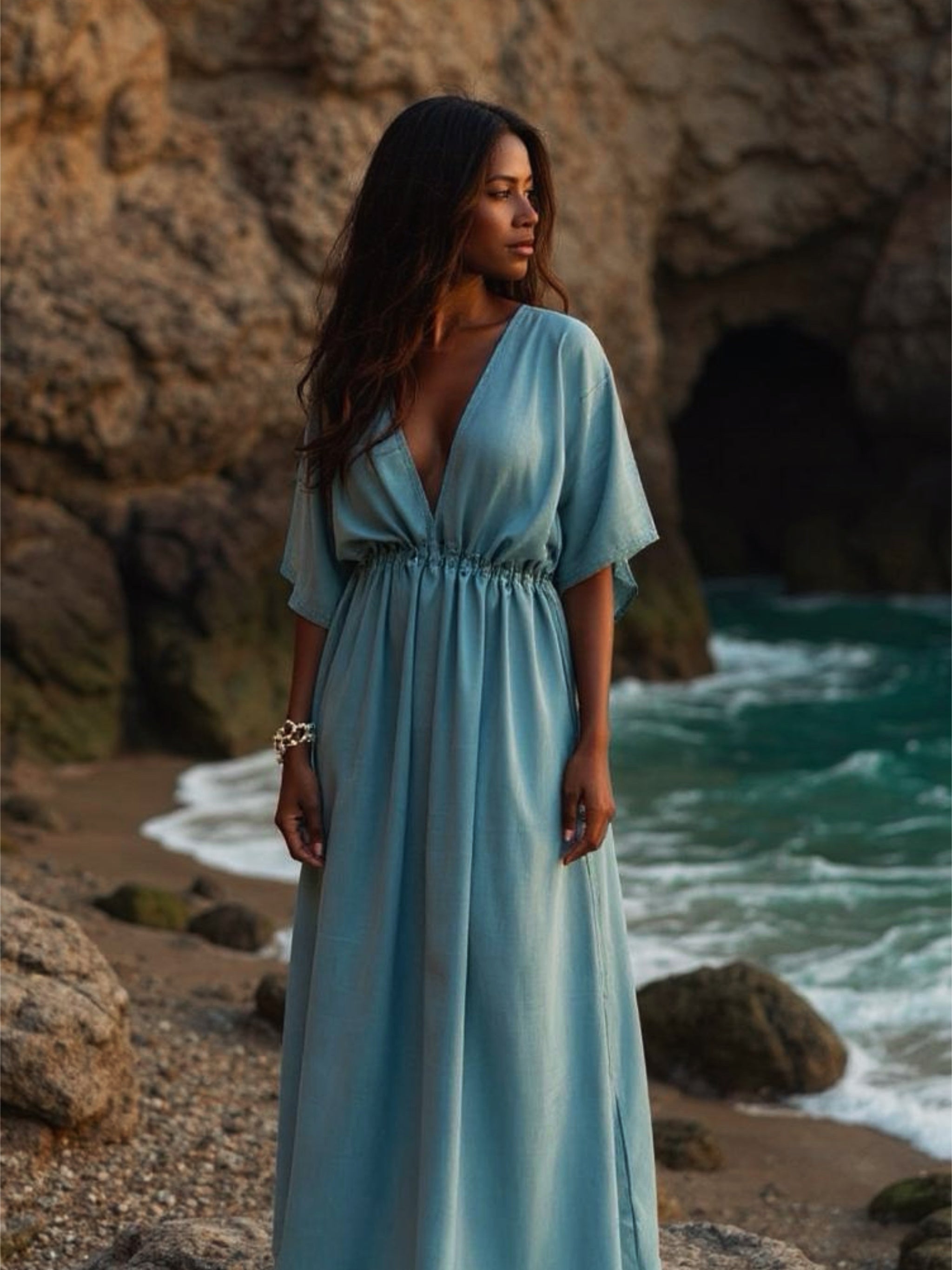 Woman wearing teal tie dye goddess kaftan maxi dress with sunglasses and straw hat on a tropical beach with ocean and cave in the background