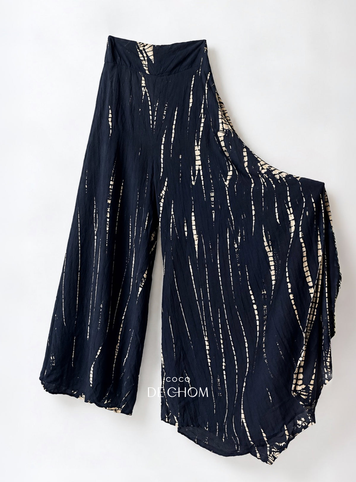 A pair of loose, wide-leg cropped tie dye pants in black with a vertical tie-dye style pattern of beige and white stripes.