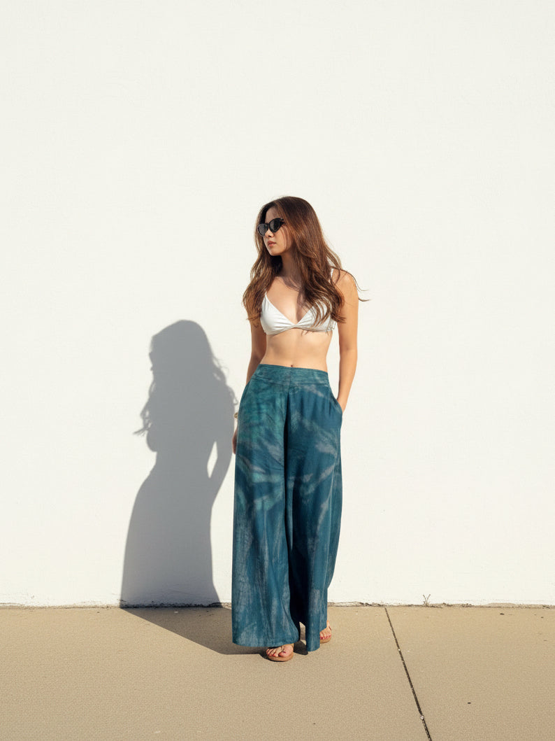 Woman wearing a white top and teal tie dye boho pants standing against a white wall with sand at the bottom, perfect beach outfit.