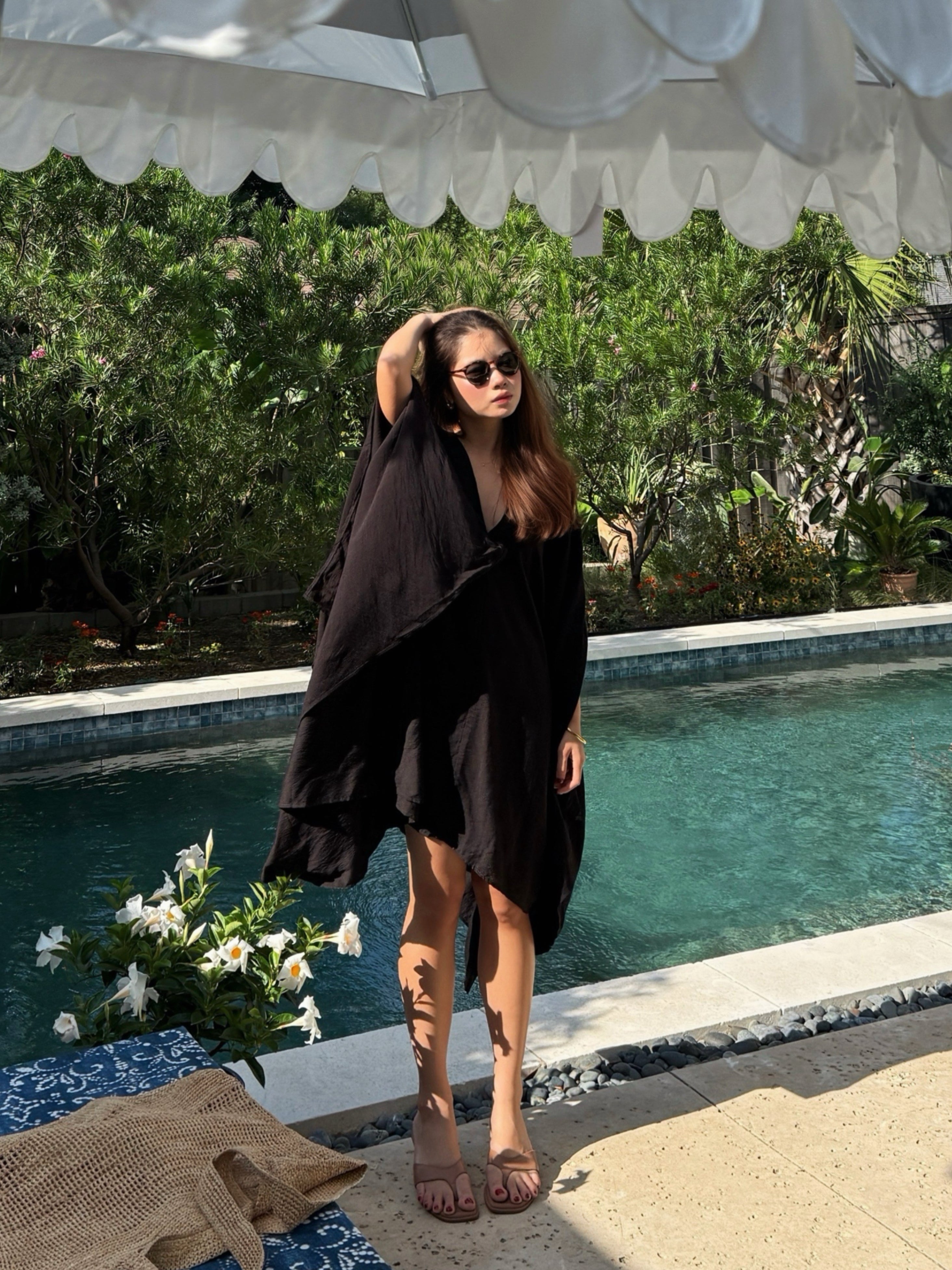 A woman wearing a Black Mini Kaftan Cover Up, oversized beach cover up and petite mini dress with deep V neckline for resort wear and poolside vacation outfits stands by a swimming pool under a white patio umbrella, with green foliage in the background.