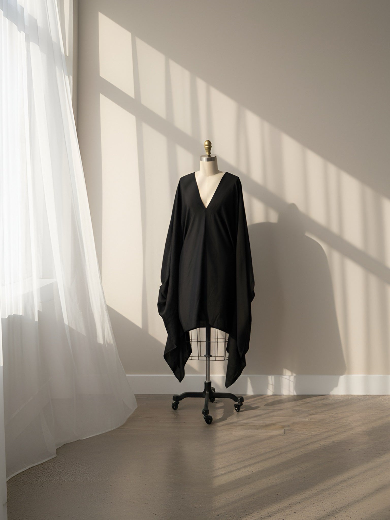 A black draped V-neck top or short kaftan displayed on a dress form mannequin in a minimalist room with warm sunlight casting window shadows on a beige wall and light wood floor.