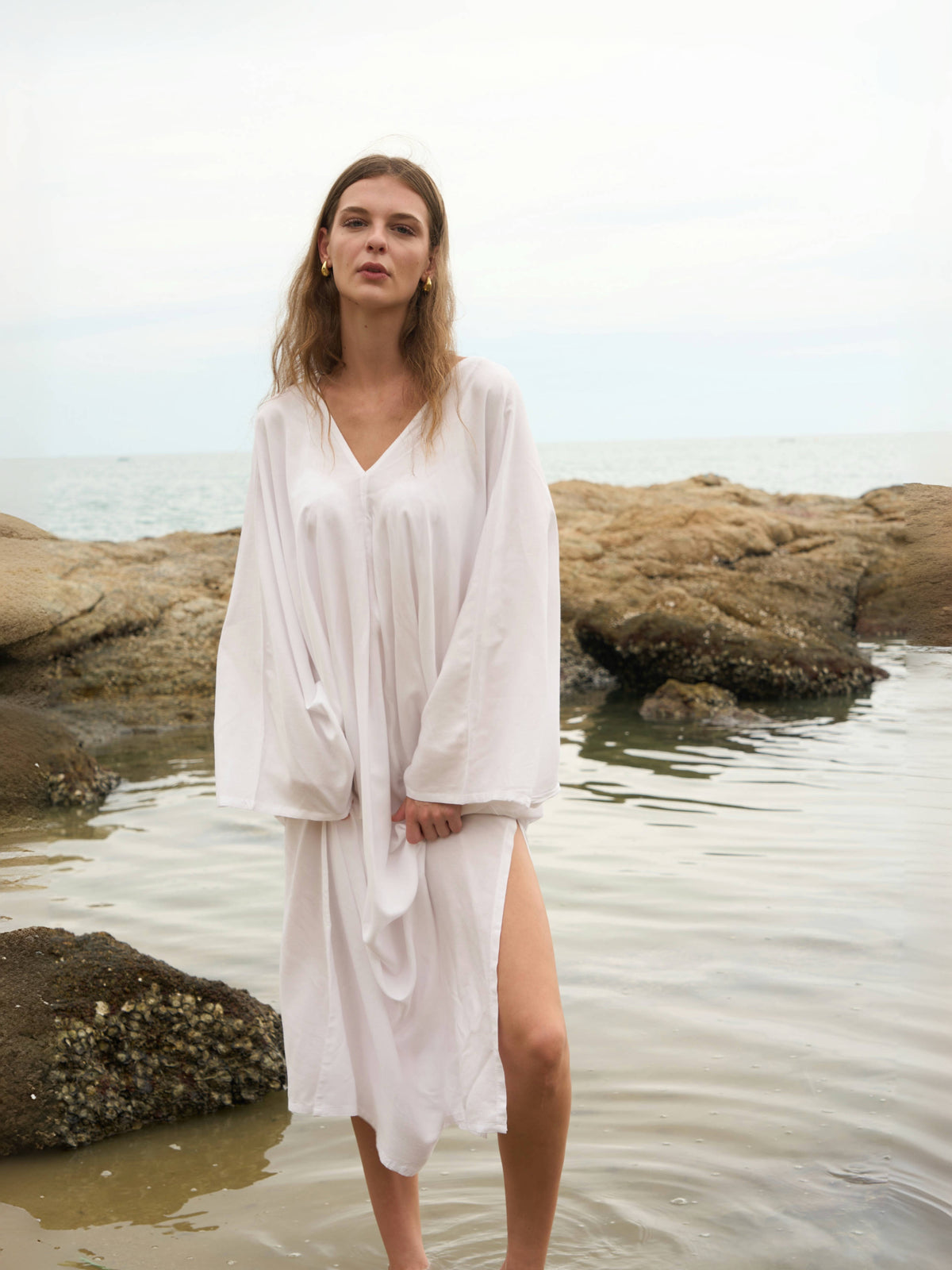 A woman wearing a white long-sleeve maxi kaftan dress stands in shallow water on a rocky beach with the ocean in the background.