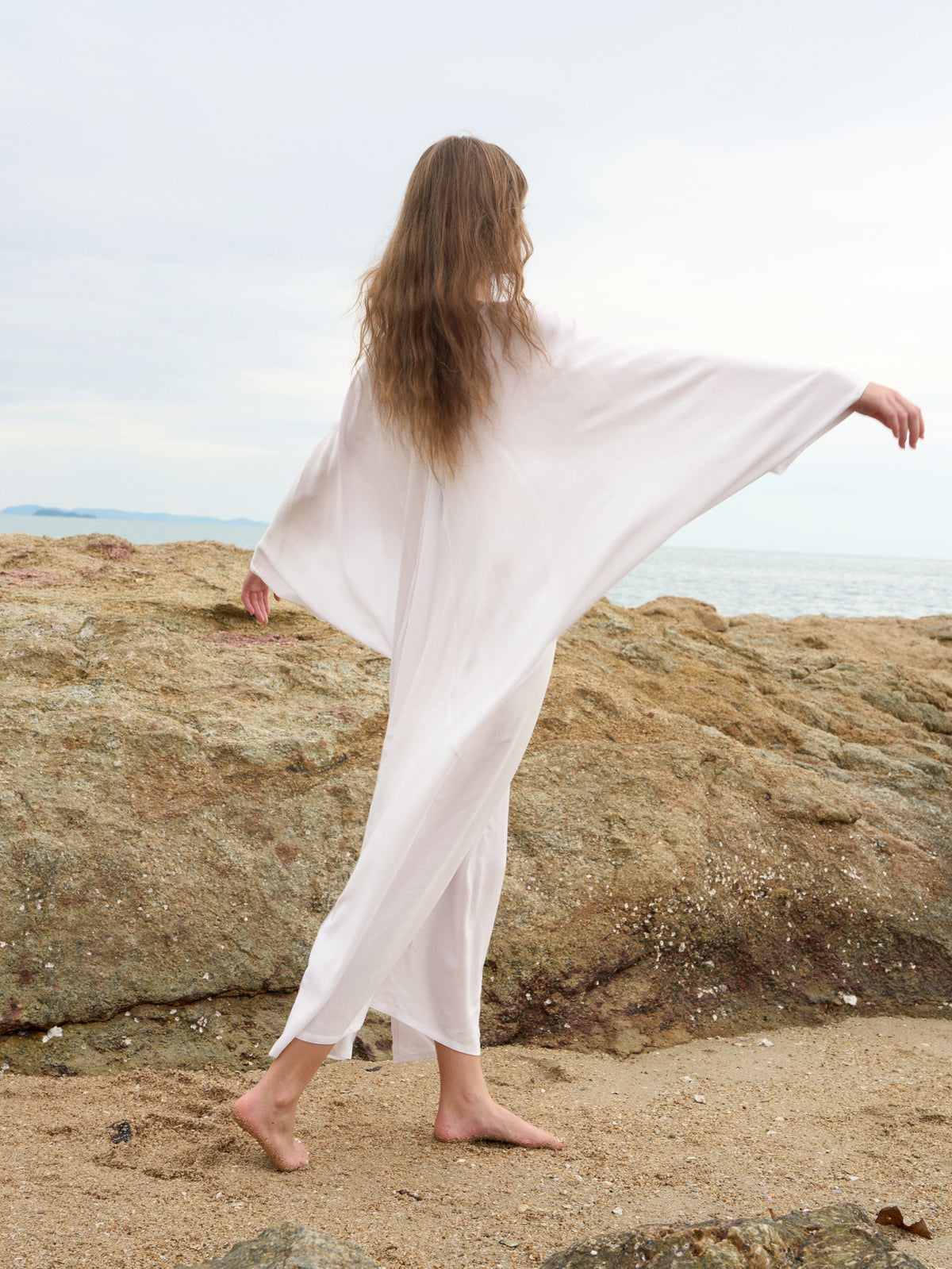 A woman with long light brown hair stands barefoot on a sandy beach with large rocks and the ocean in the background, wearing a flowing, loose white cover-up dress or kaftan with wide sleeves.