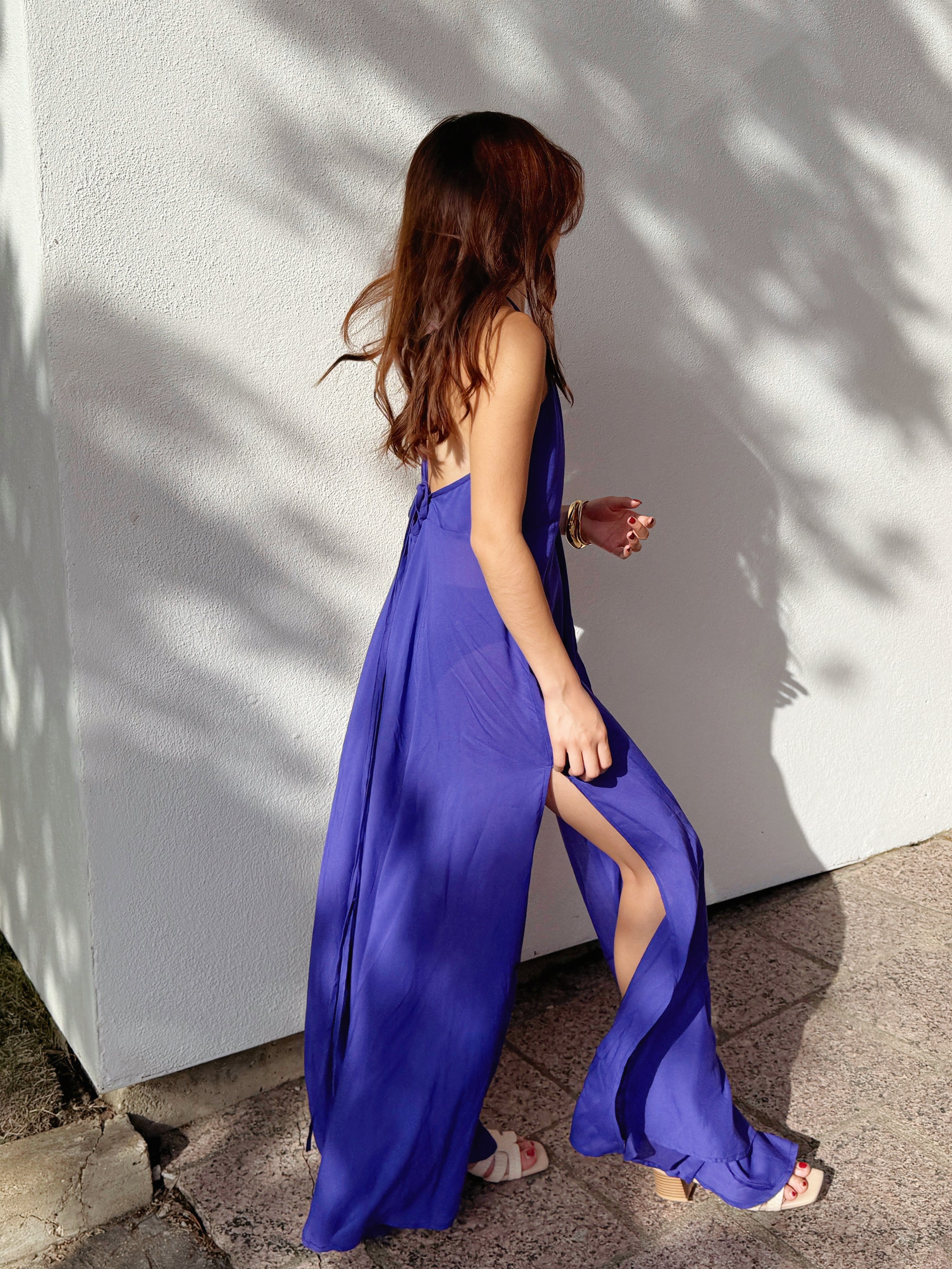 Woman wearing a flowy, hand-dyed purple halter maxi dress with an open back and high slit, walking confidently against a white wall with shadows.