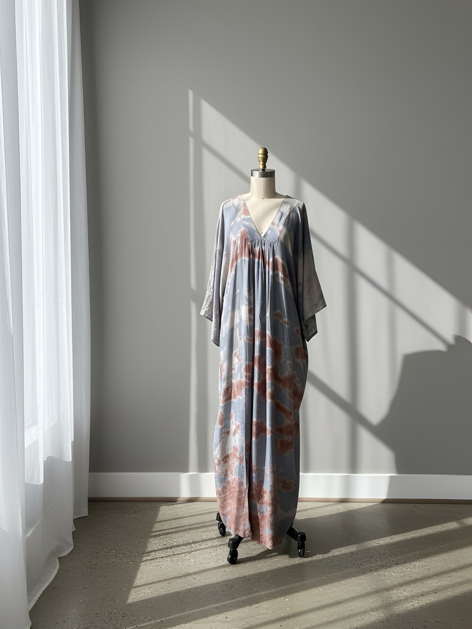 Mannequin wearing a tie-dye kaftan dress in a sunlit room with shadows on the wall.