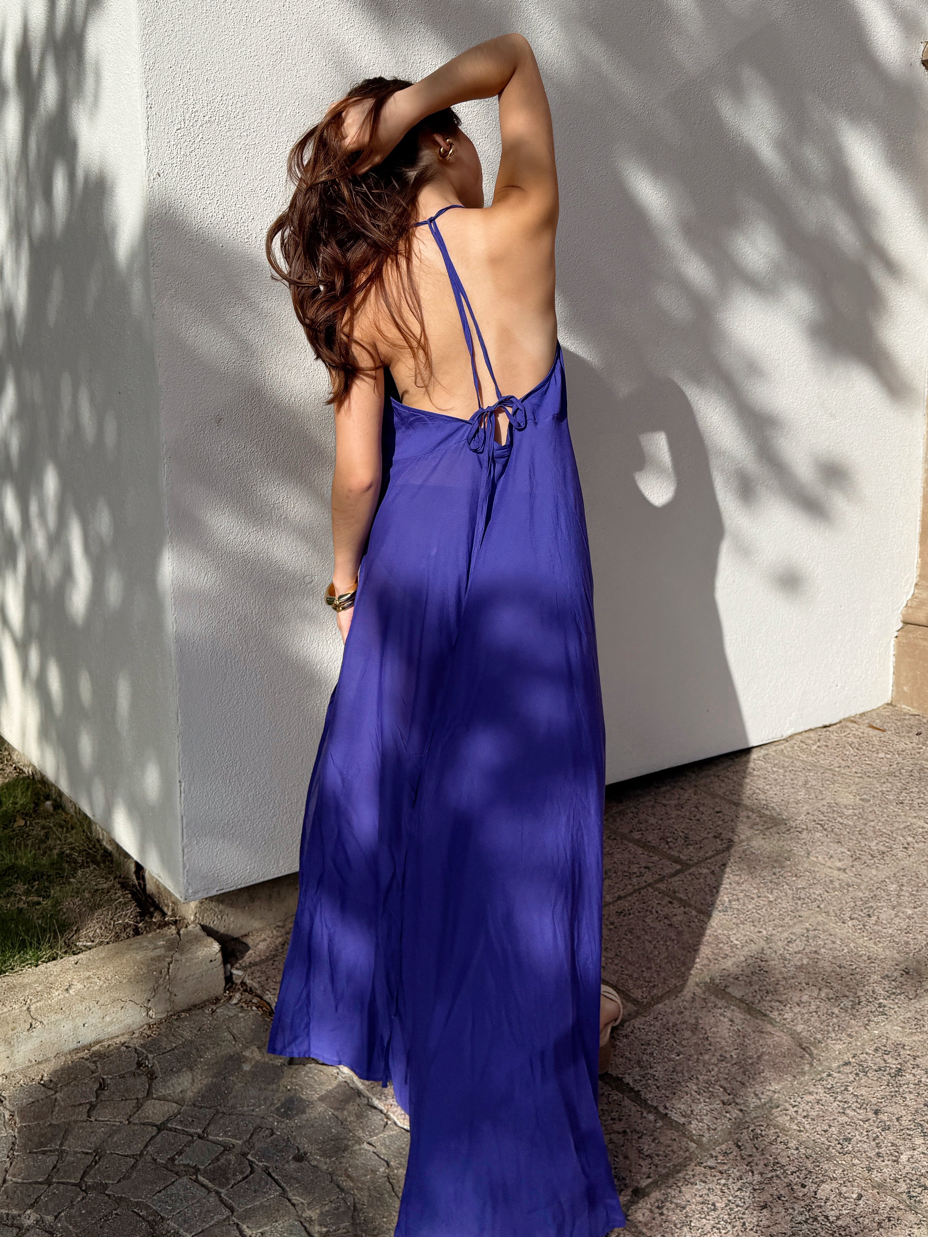 Woman wearing a flowy, hand-dyed persian indigo halter maxi dress It features an open-back design with thin, adjustable halter-style straps that tie around the neck and crisscross down the back. A second set of long ties wraps around the mid-back and waist, allowing for a customizable fit and added shaping.