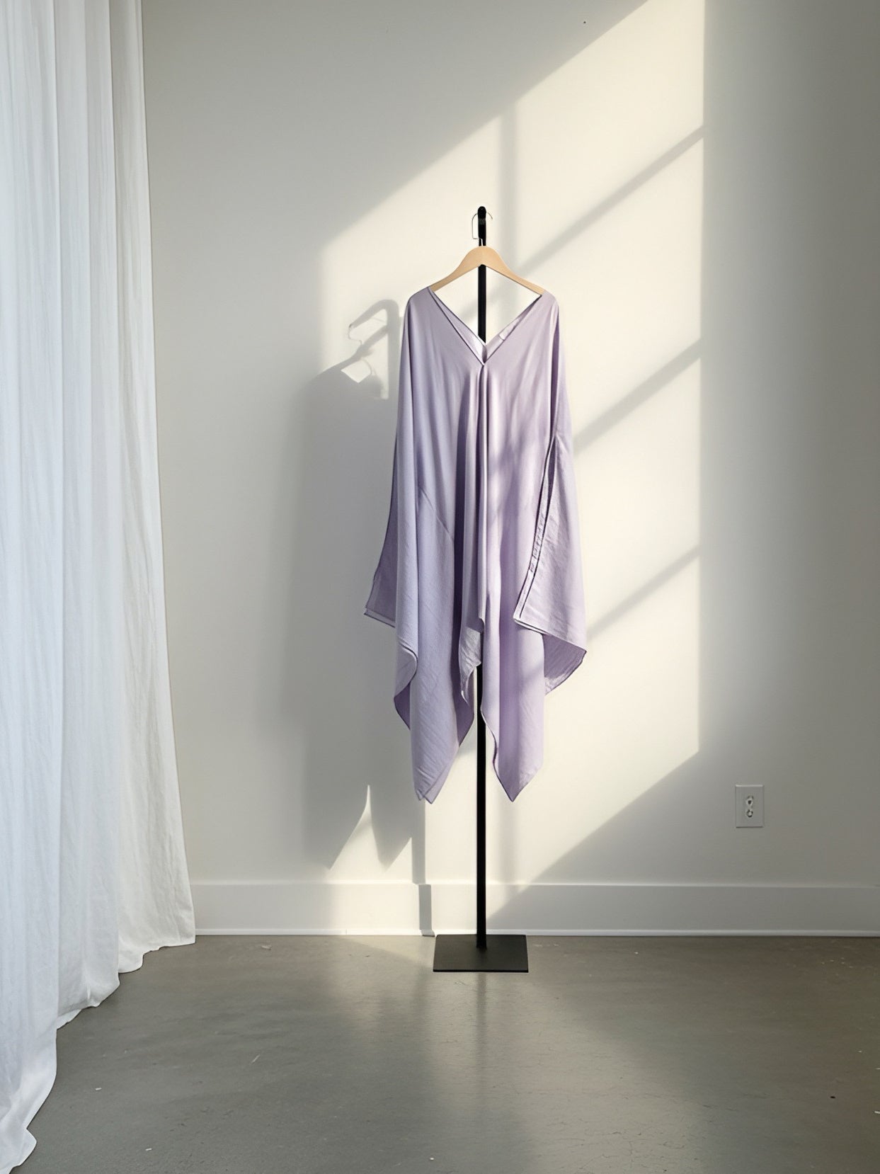 A lavender purple kaftan-style tunic or mini kaftan dress top with a V-neck and wide sleeves, hanging on a black clothing stand against a white wall with soft window shadows.
