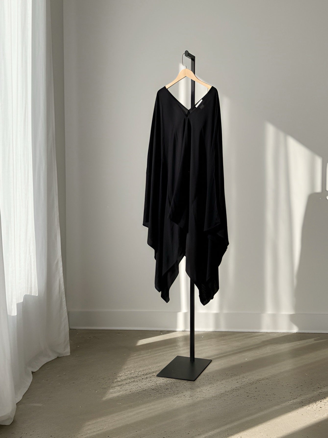 A black draped asymmetrical tunic top or kaftan dress displayed on a black stand with a wooden hanger, set against a minimalist gray wall with soft sunlight and shadows. Black Mini Kaftan Cover Up, oversized beach cover up and petite mini dress with deep V neckline for resort wear