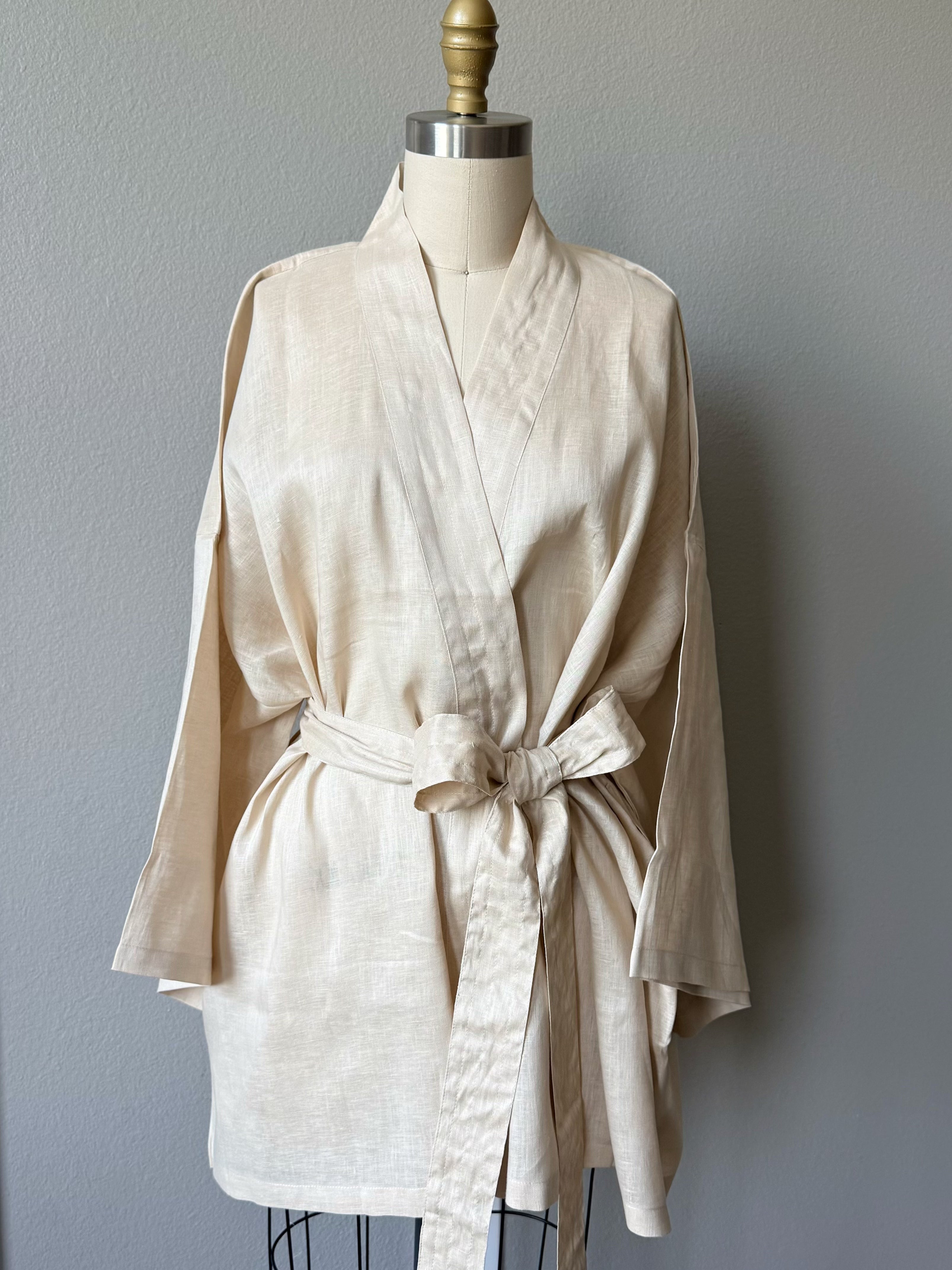 A cream linen kimono-style robe with a tie belt, displayed on a dress form mannequin against a gray wall.