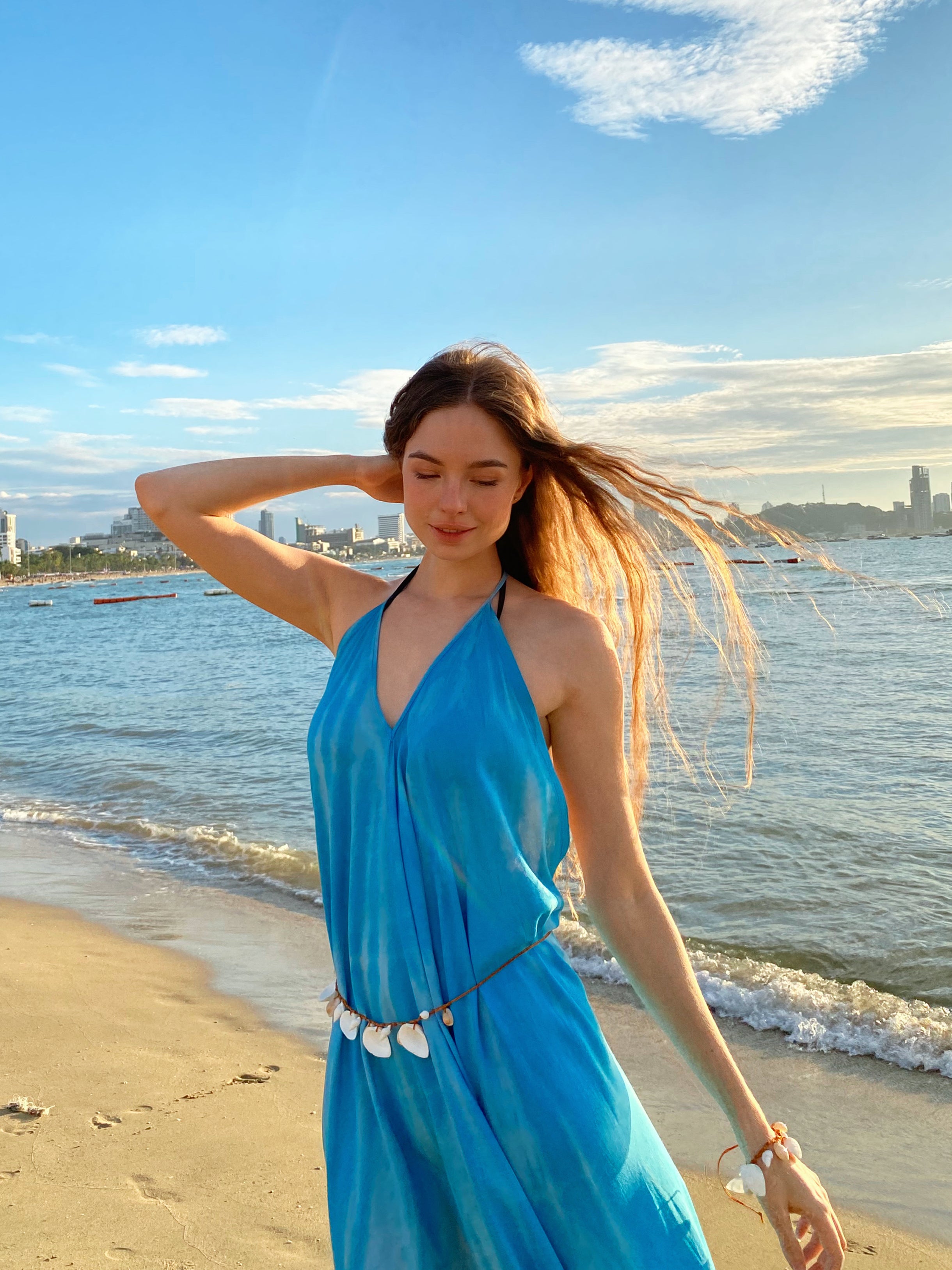 Woman in a blue maxi dress standing on a beach with a city skyline in the background, beach dress in blue perfect for vacatioon