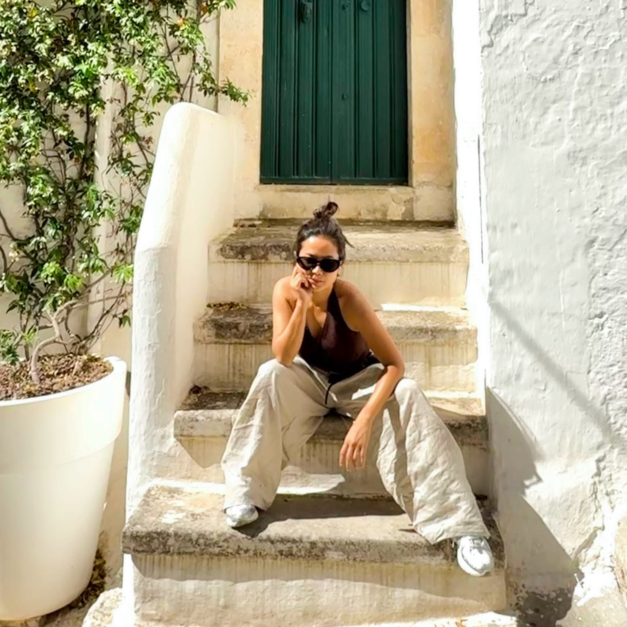 Woman sitting on stone steps in  natural linen wide leg pants in a sunlit alleyway with white walls and green door.