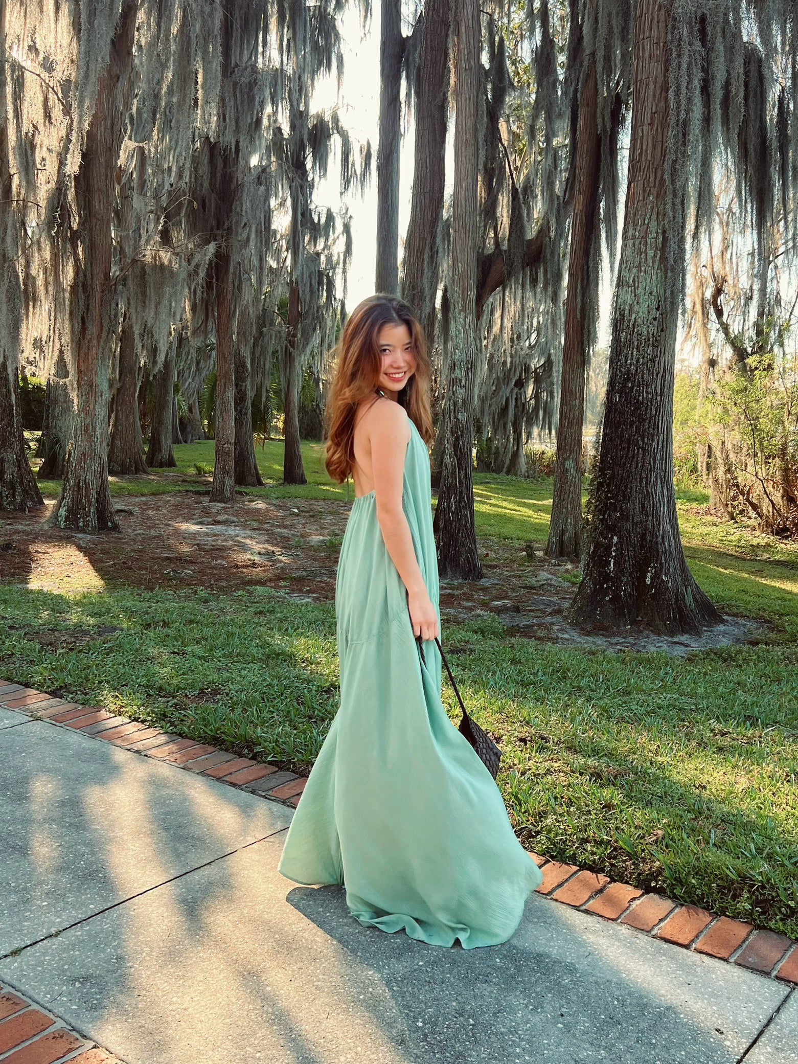 Woman in a sage green maxi dress with cape detail standing in a sunlit hallway, a handmade resort dress and beach wedding guest style by COCO DE CHOM