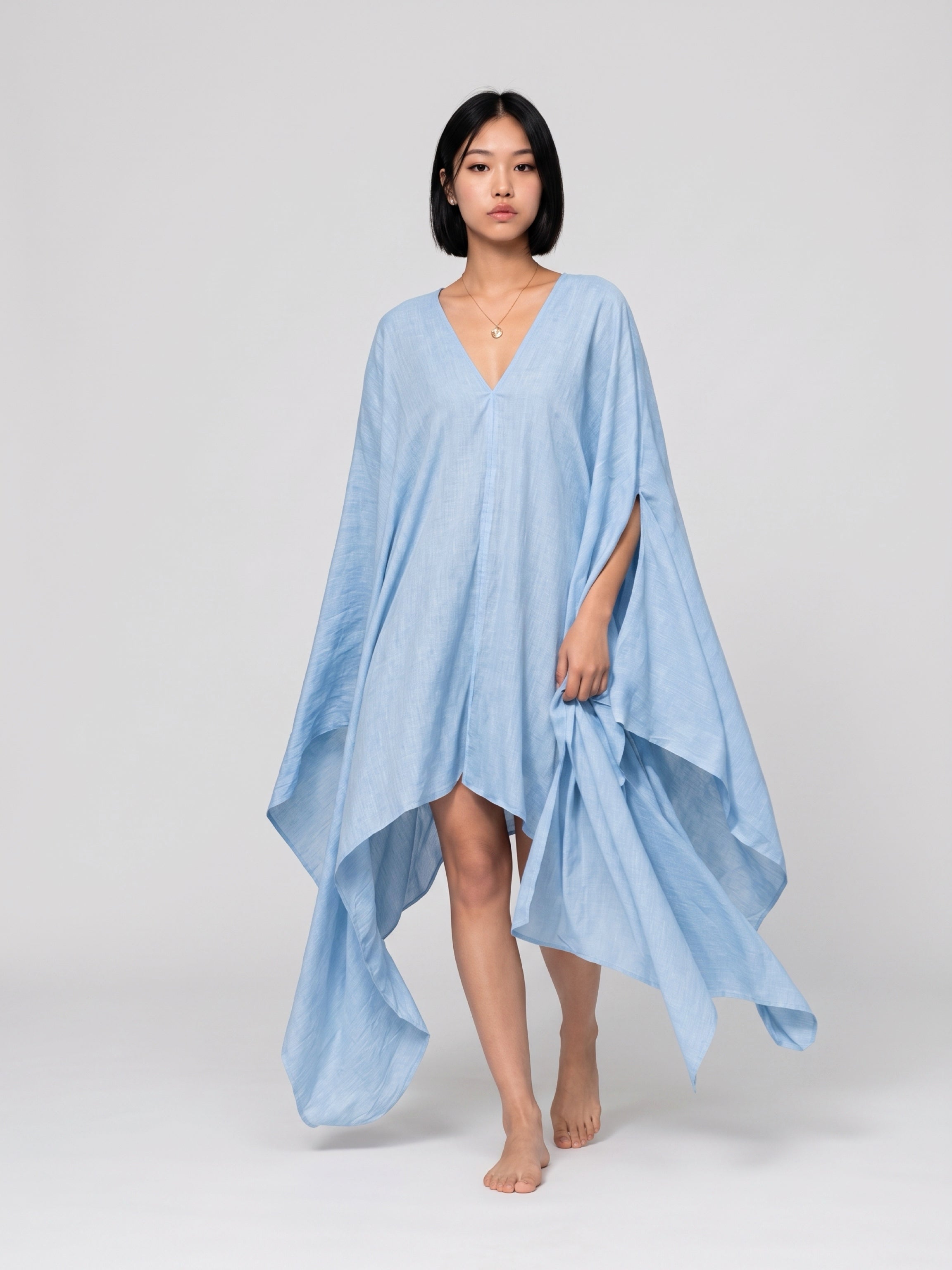 A woman modeling a light blue kaftan dress with a V-neckline and wide draped sleeves, standing against a neutral light gray background. Sky blue Soleil Mini Kaftan Cover Up, lightweight mini kaftan dress and beach cover up styled as a resort top for summer vacation