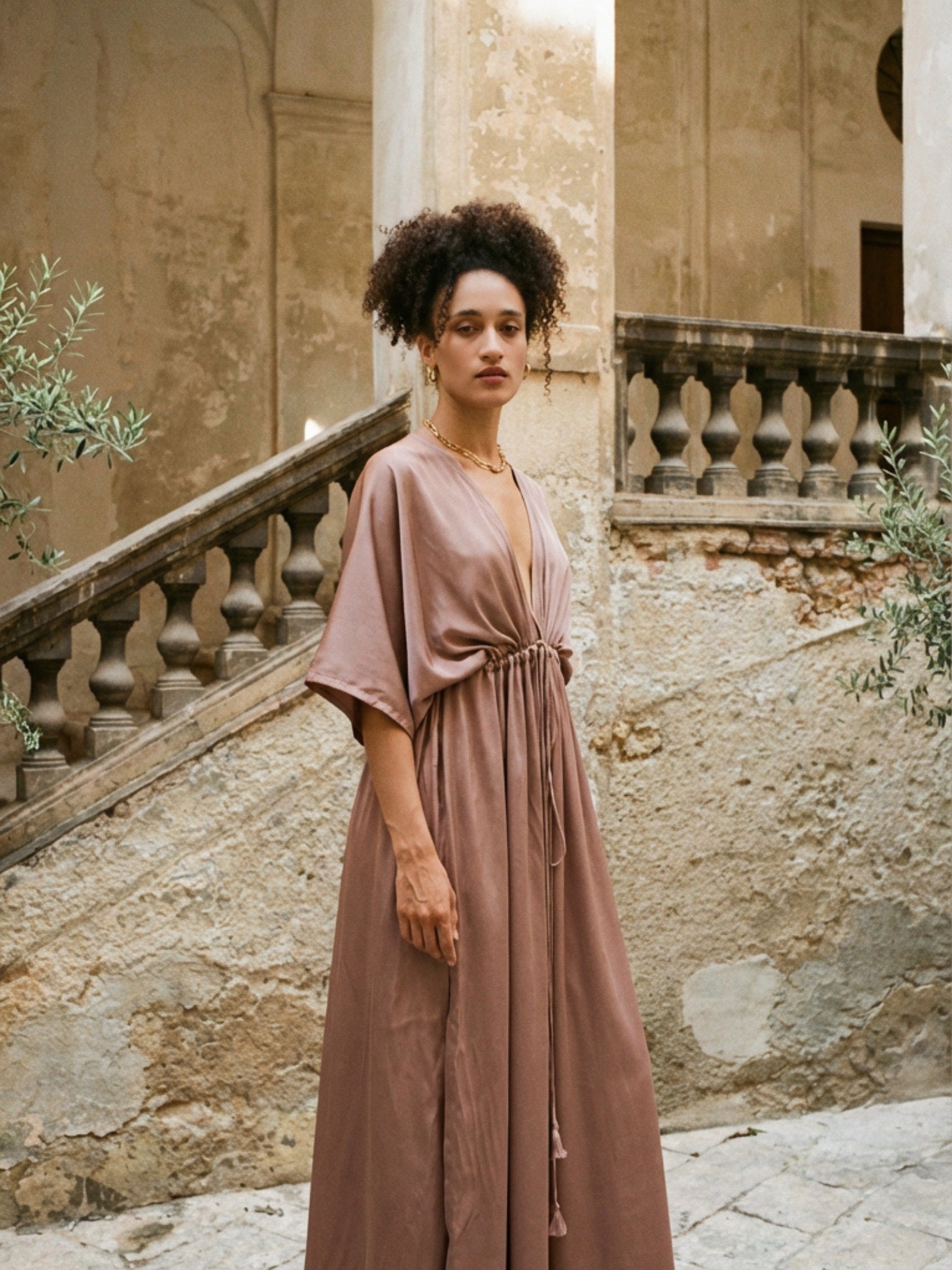 Woman wearing a mocha rose kaftan maxi dress with deep V-neck and self-tie waist, styled as resort wear and a beach wedding guest dress by COCO DE CHOM