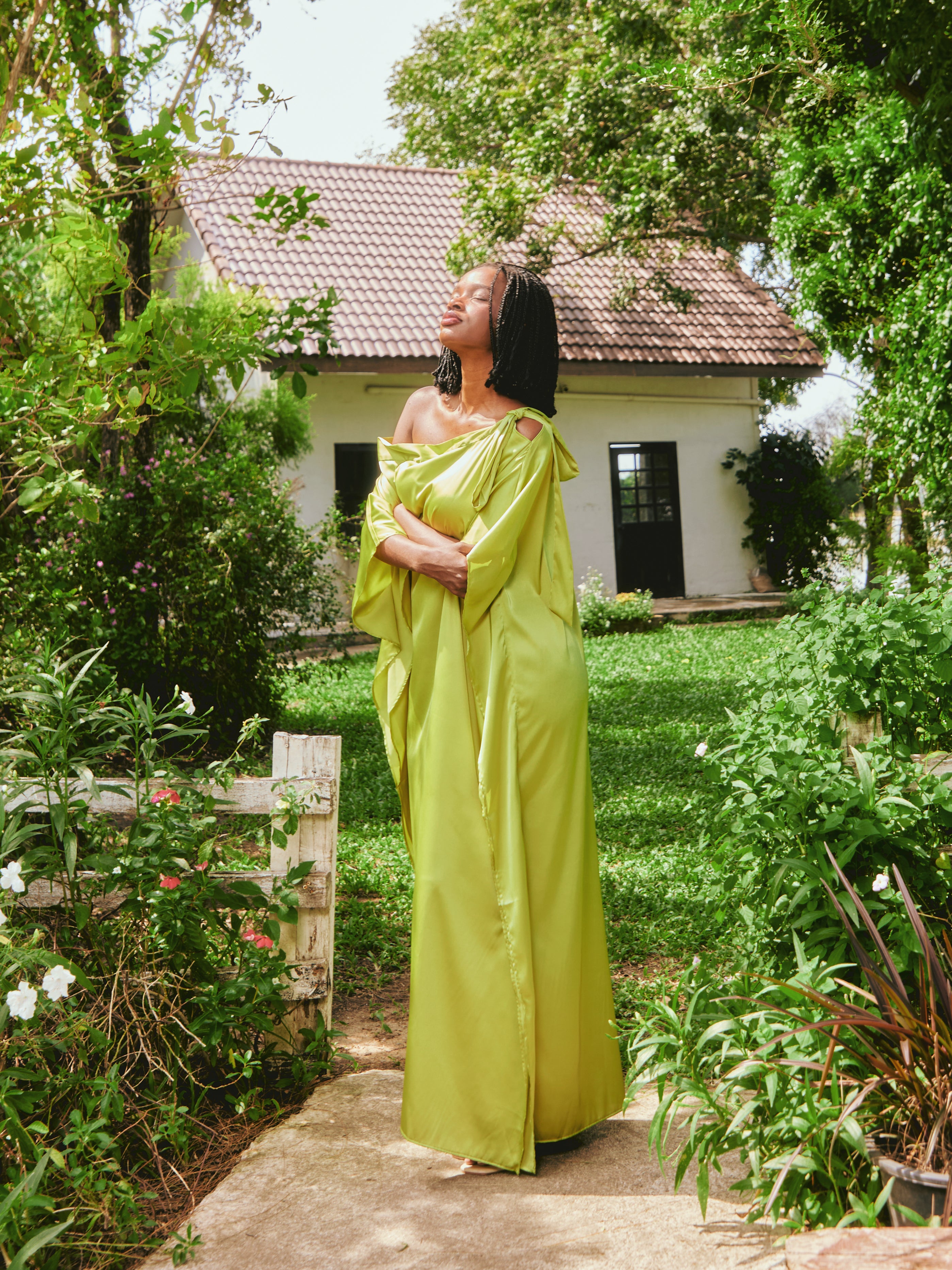 Woman in a lemon green kaftan dress standing on grass with a house and tree in the background. The Helena Lemon Green Kaftan by Coco De Chom is an off-shoulder satin resort dress with a free-flowing silhouette, tie details, and silky eco-friendly fabric — perfect for beach vacations, resort wear, and effortless boho luxury.
