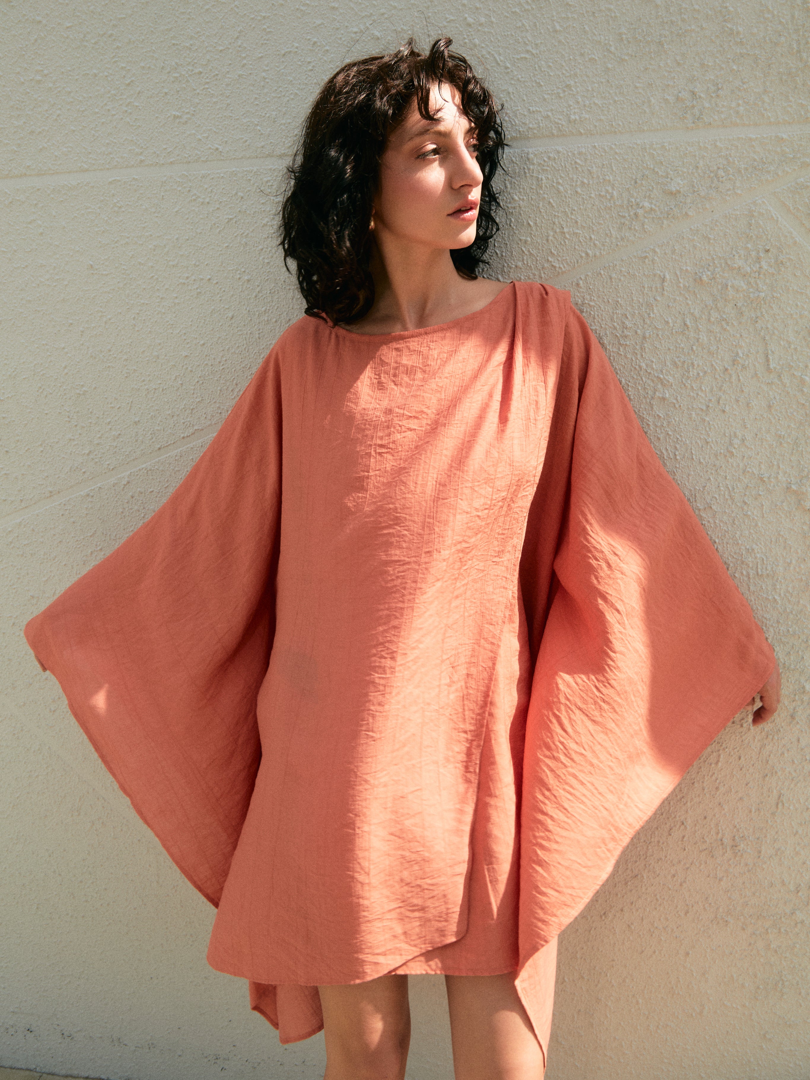 Woman wearing a coral mini kaftan dress against a light background -flowy oversized tunic for beach and pool, airy drape with long sleeves and high low hem