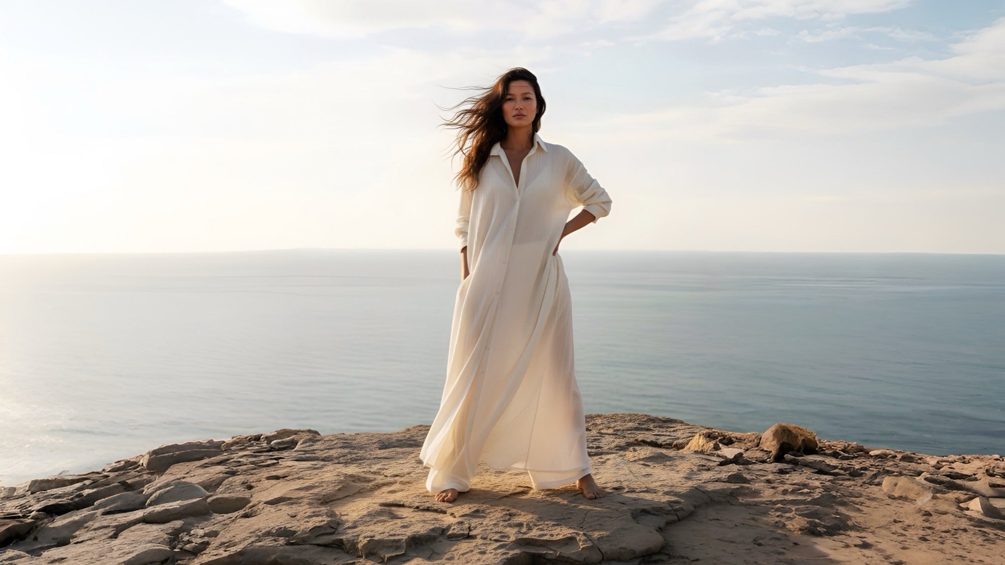 Woman in a white cotton shirt dress standing on a rocky beach with ocean and sky in the background