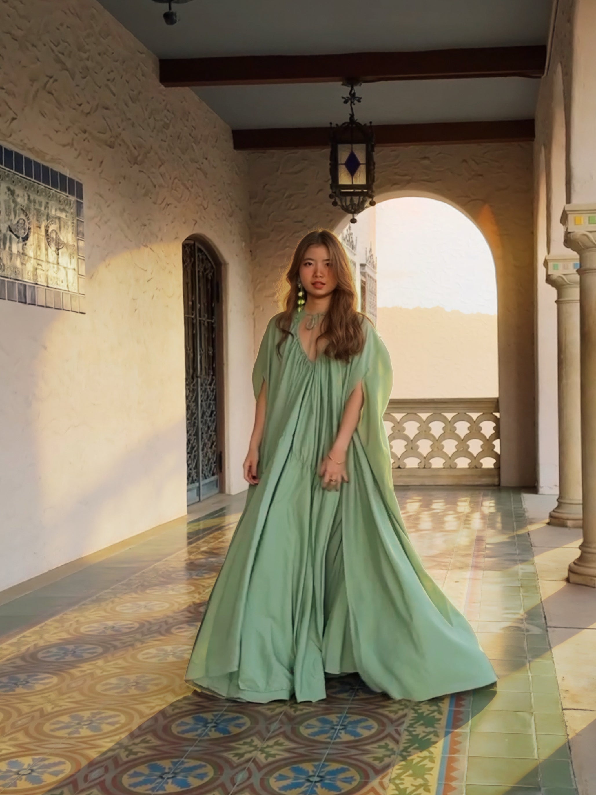 Woman in a sage green maxi dress with cape detail standing in a sunlit hallway, a handmade resort dress and beach wedding guest style by COCO DE CHOM