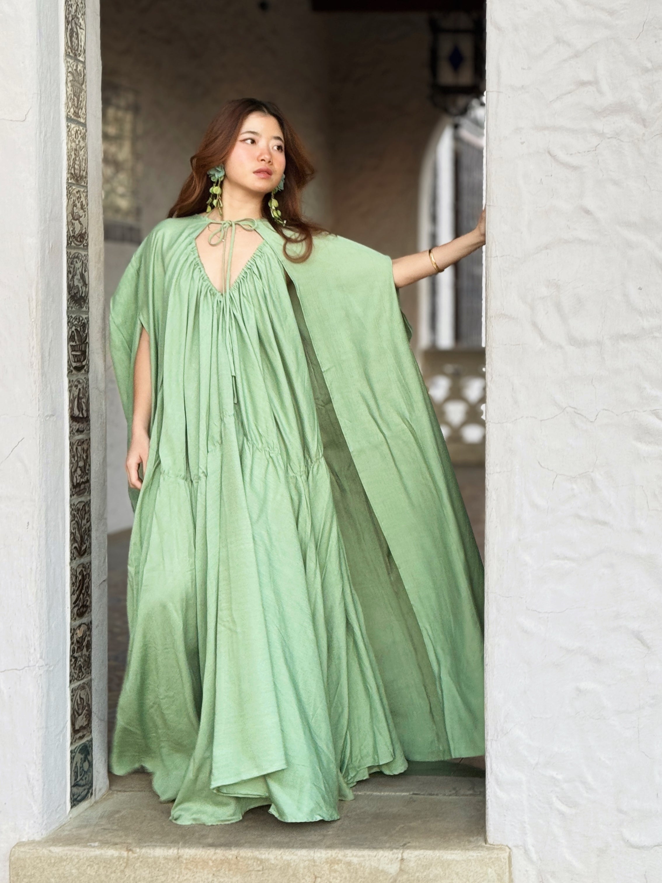 Woman in a sage green maxi dress with cape detail standing in a sunlit hallway, a handmade resort dress and beach wedding guest style by COCO DE CHOM
