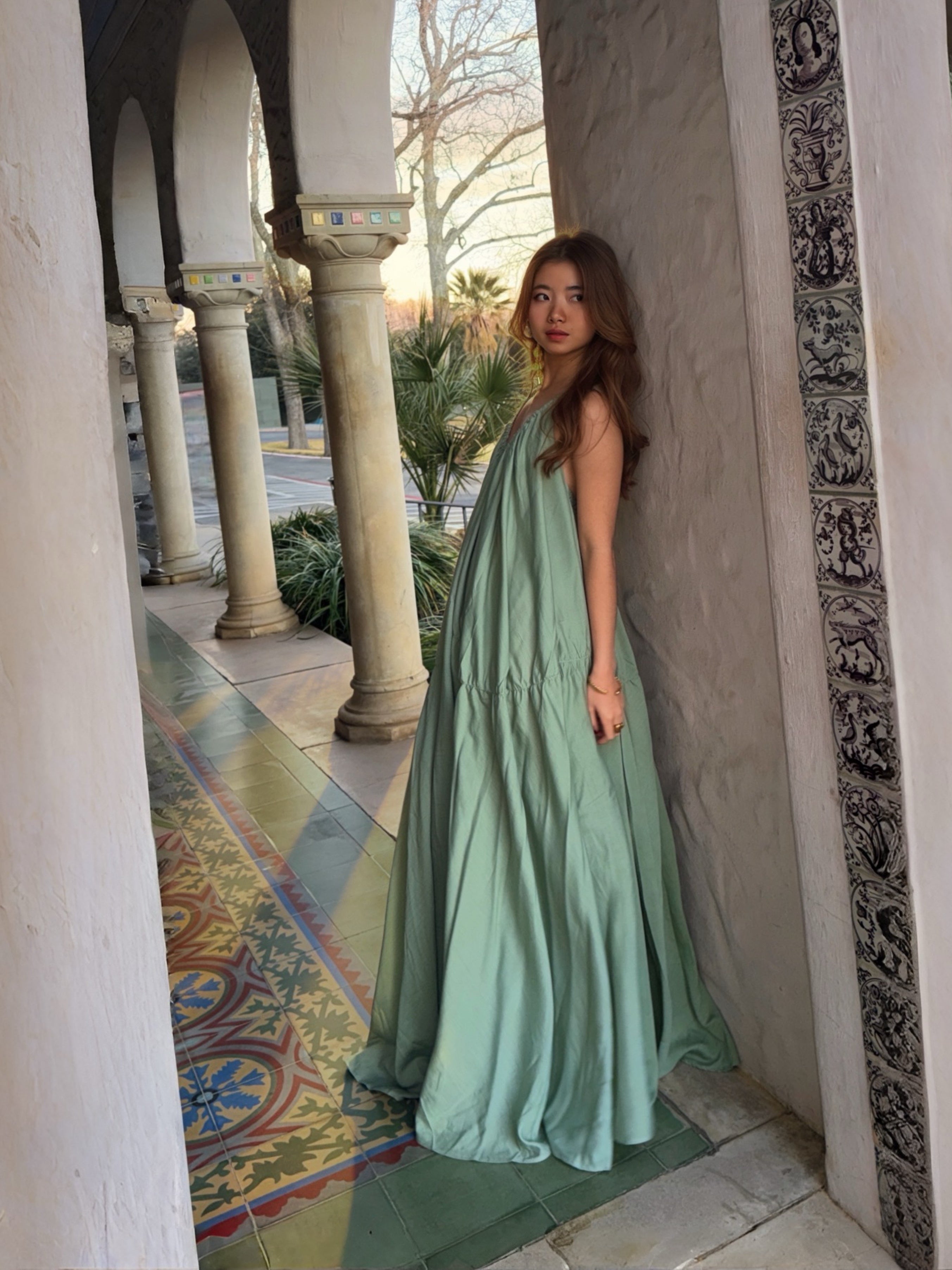 Woman in a sage green maxi dress with cape detail standing in a sunlit hallway, a handmade resort dress and beach wedding guest style by COCO DE CHOM
