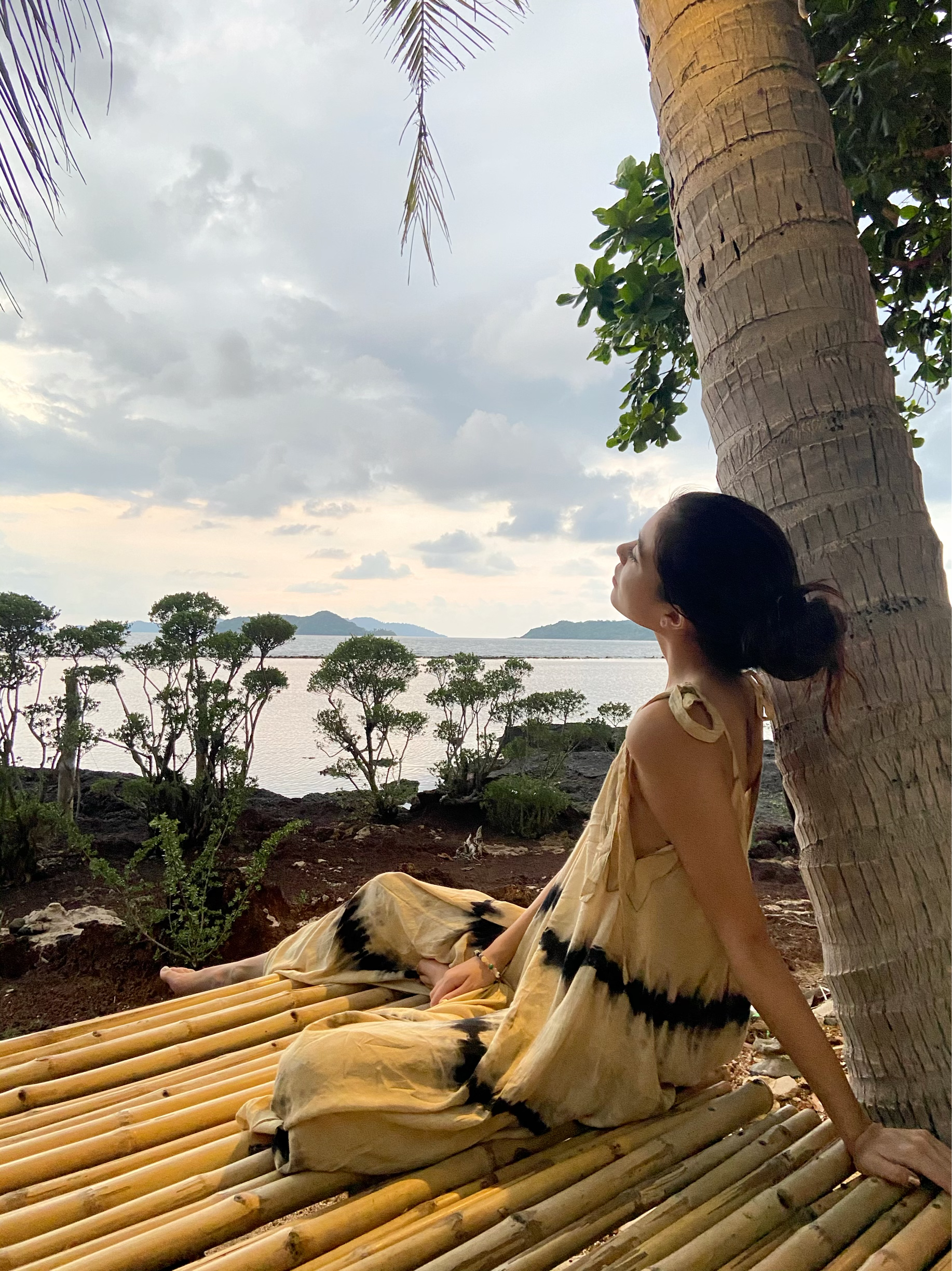 Woman in tie dye jumpsuit with an oversized fit and back-V silhouette, perfect for yoga, beach cover-up, or casual day-to-night wear by Coco De Chom standing on a sandy beach with ocean and rocks in the background.
