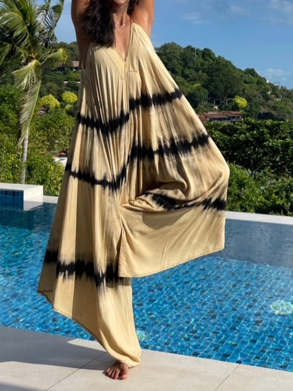 Woman in tie dye jumpsuit with an oversized fit and back-V silhouette, perfect for yoga, beach cover-up, or casual day-to-night wear by Coco De Chom standing on a sandy beach with ocean and rocks in the background.