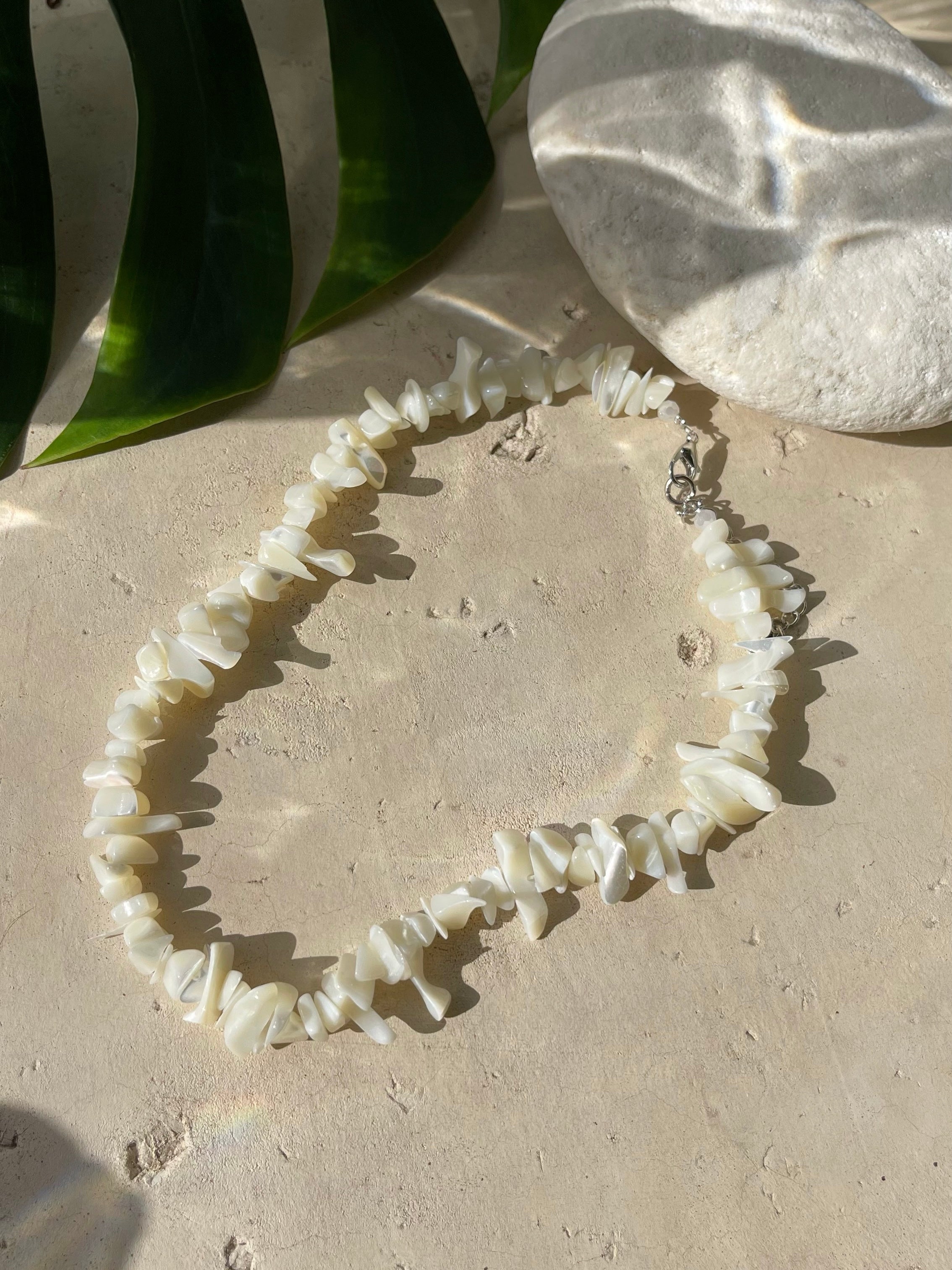 A white mother of pearl choker necklace laid out on a sandy surface with a leaf in the background, representing a beach setting.